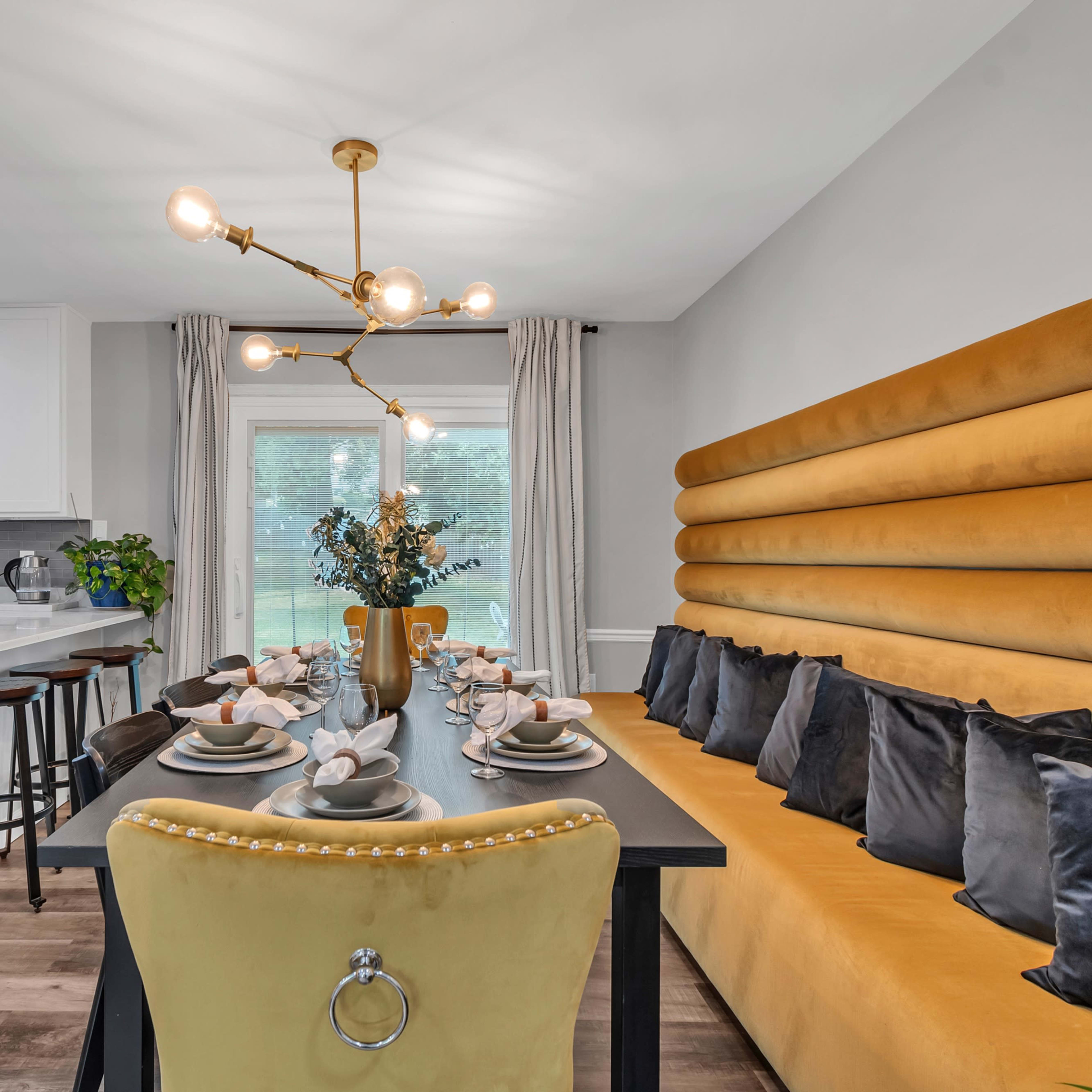 A modern dining area featuring a long table set for a meal, surrounded by a yellow upholstered banquette and stylish chairs.