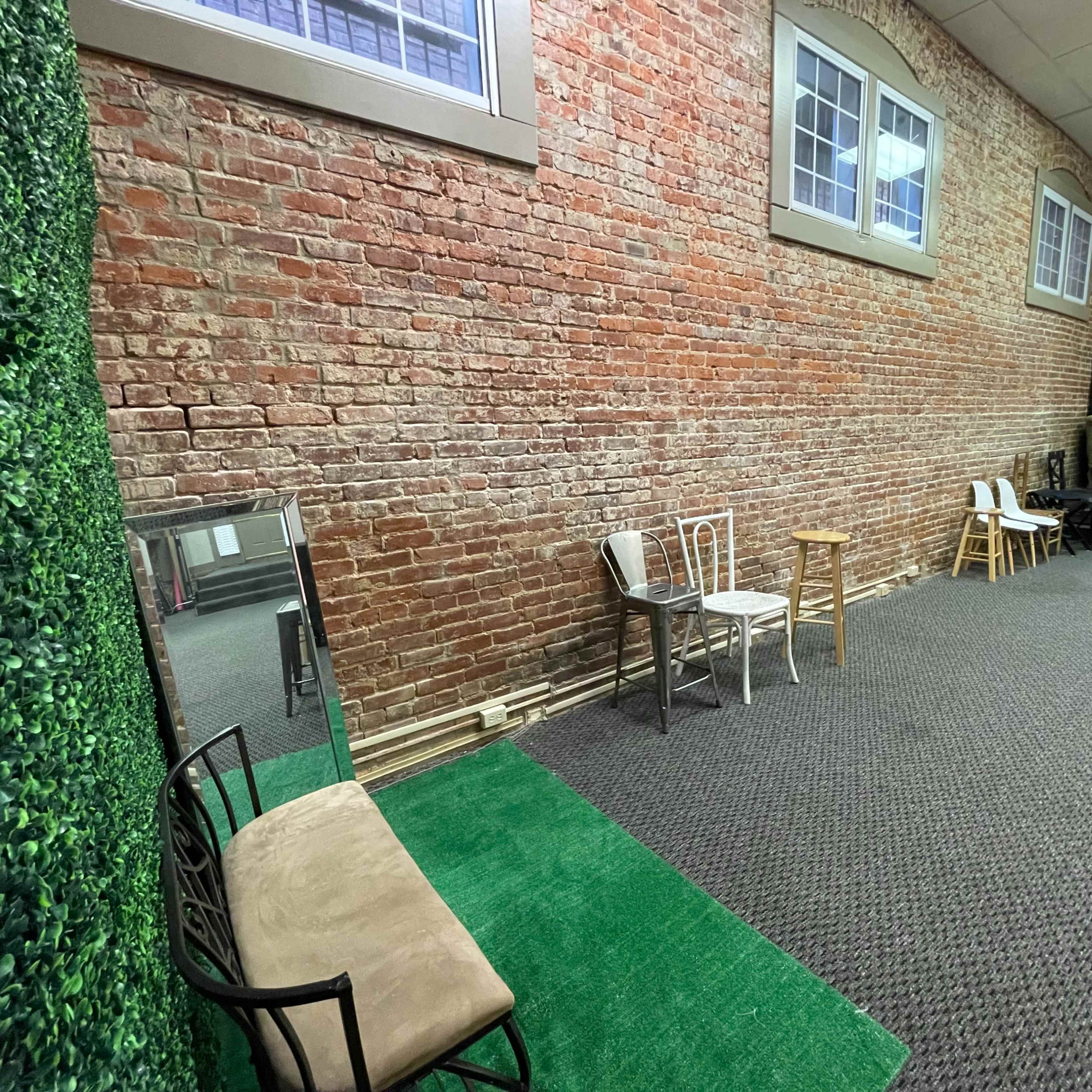 A room with exposed brick walls, two metal chairs, a small wooden stool, and a green rug, accompanied by a mirror and a couch in the background.