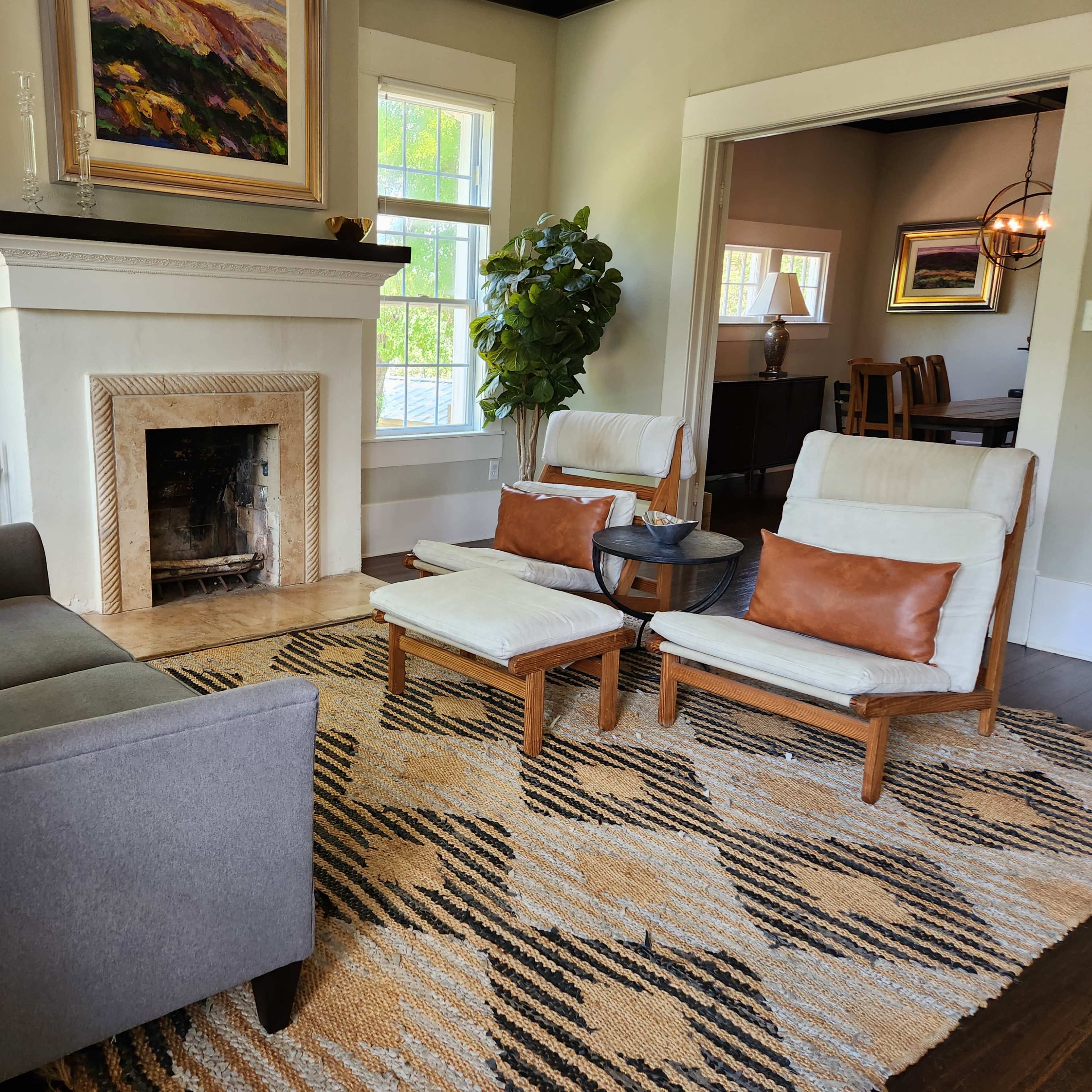 A living room with a fireplace, a gray sofa, two white chairs with brown cushions, and a large area rug.