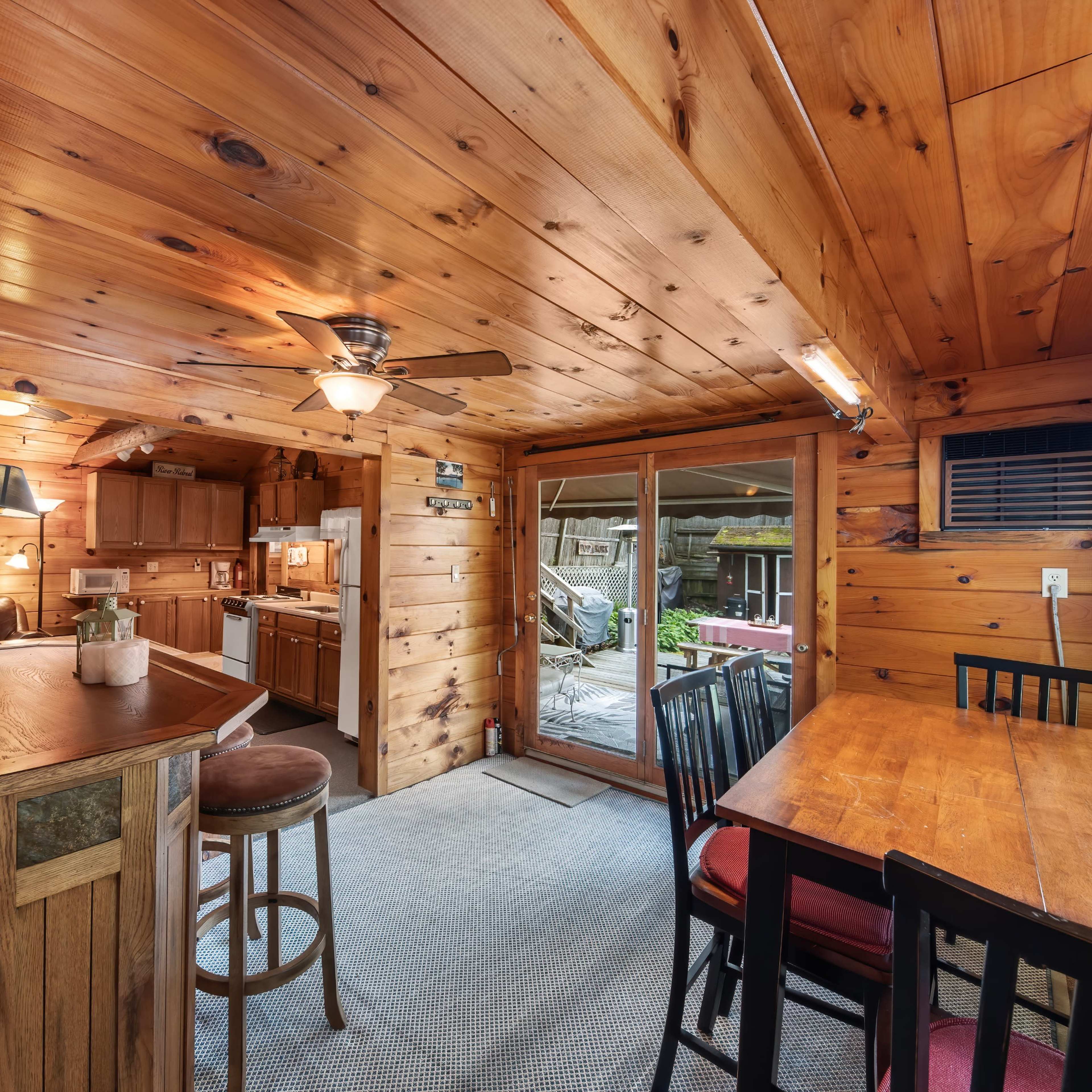 A wooden interior cabin featuring a kitchen area with a bar, dining table, and large sliding doors that open to an outdoor space.