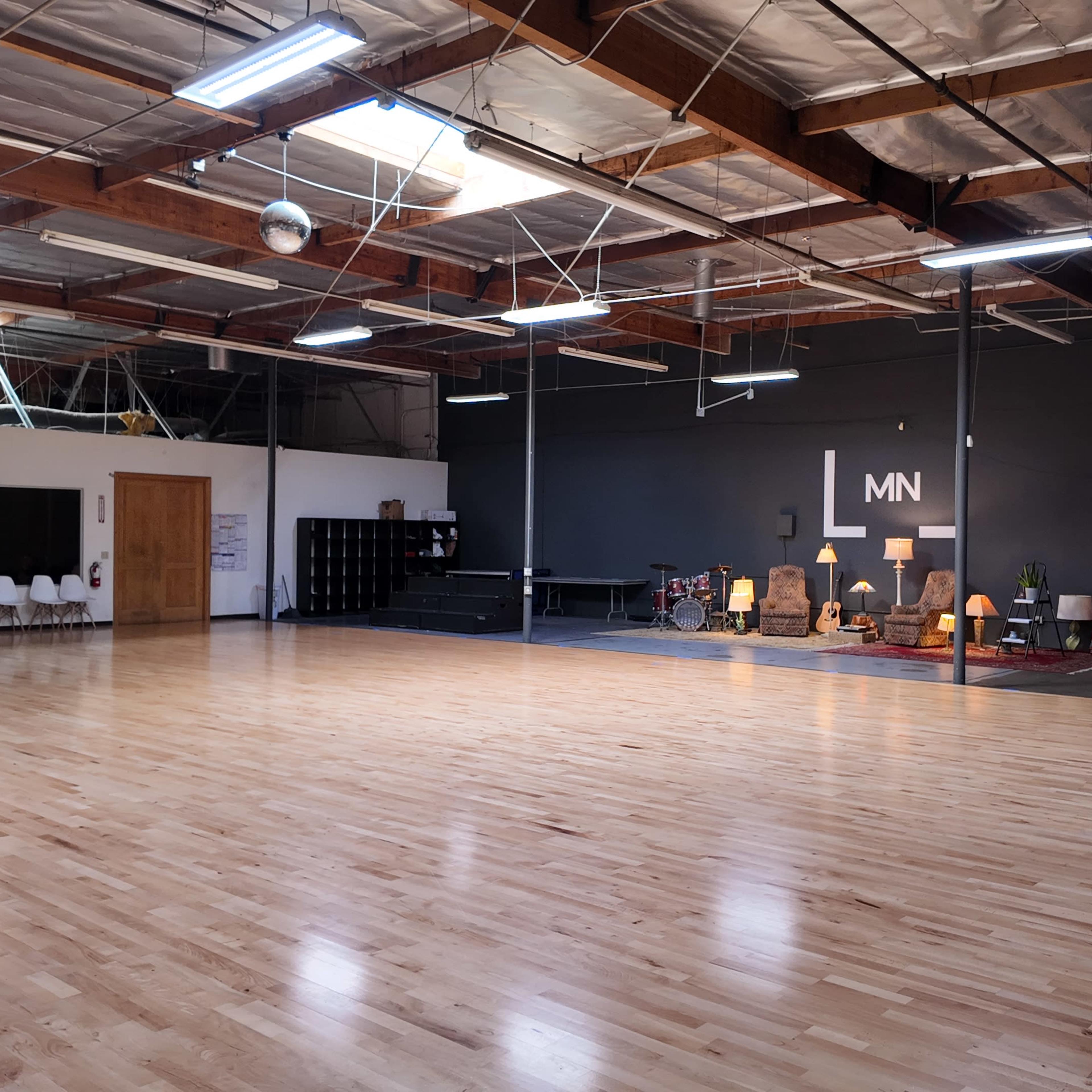 A spacious dance studio features polished wooden floors, high ceilings, and various music equipment along one wall.