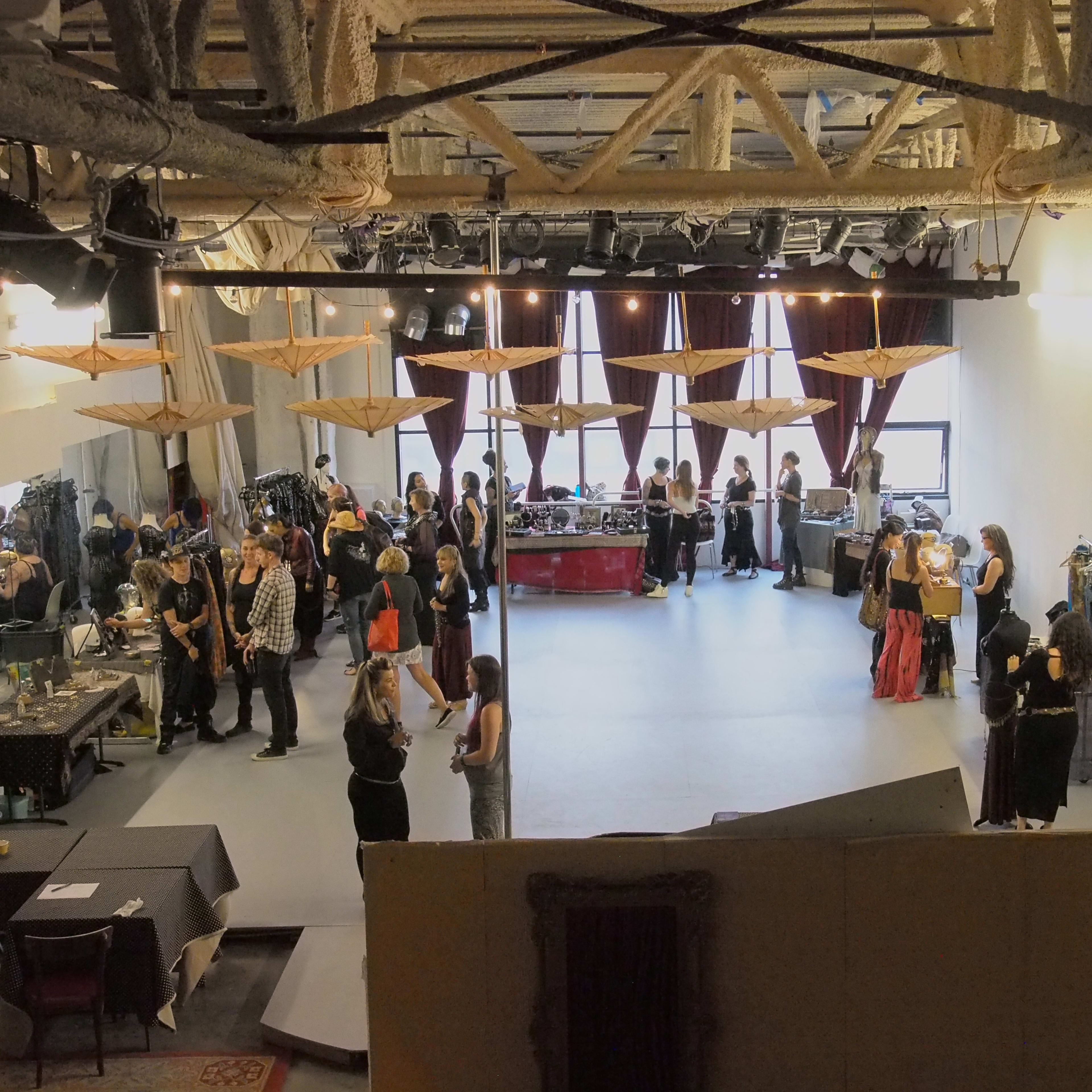 A large studio space is filled with people socializing and preparing for an event, with garments and props arranged on tables around the room.