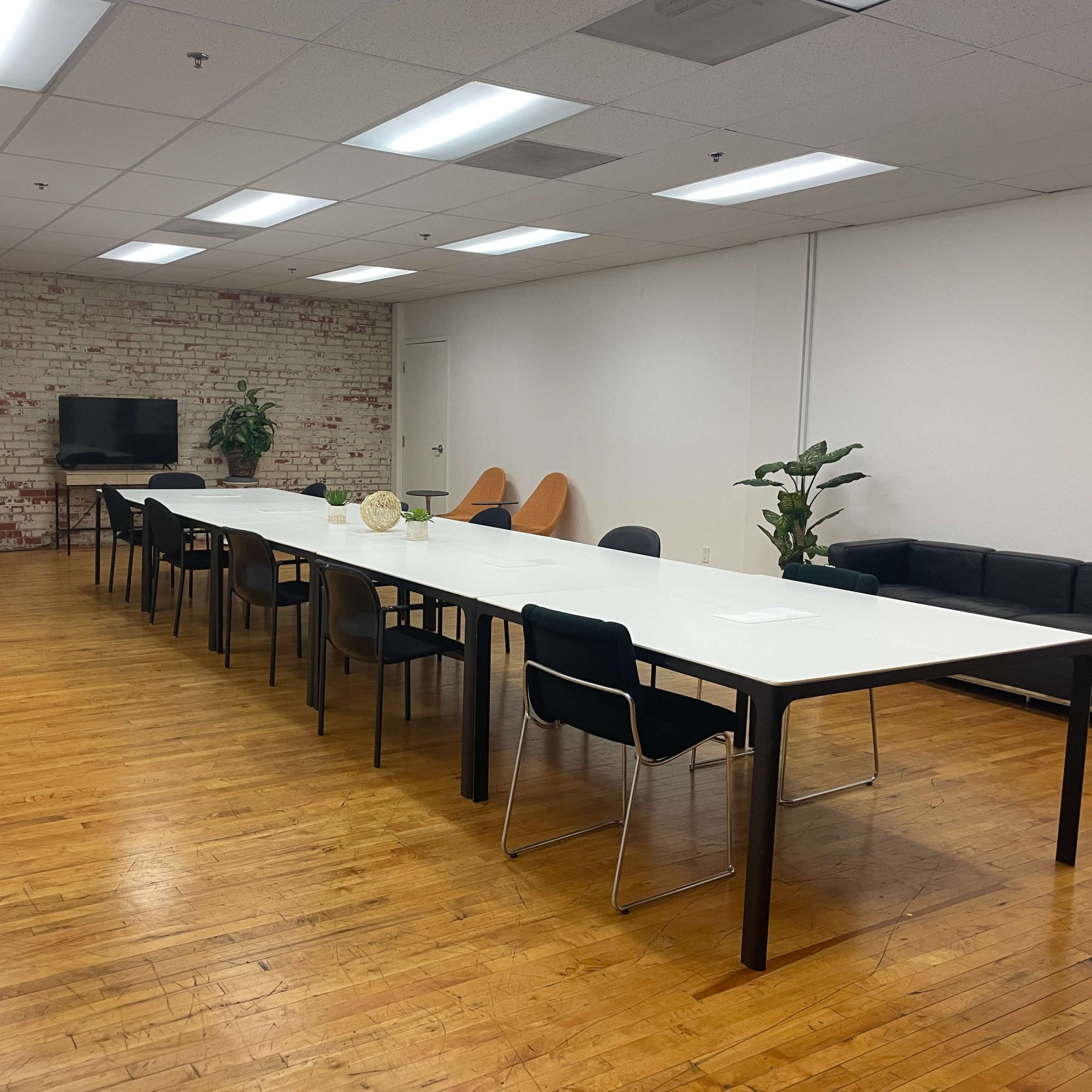 A spacious meeting room features a long white table surrounded by black and orange chairs, with a couch, television, and plants along the walls.