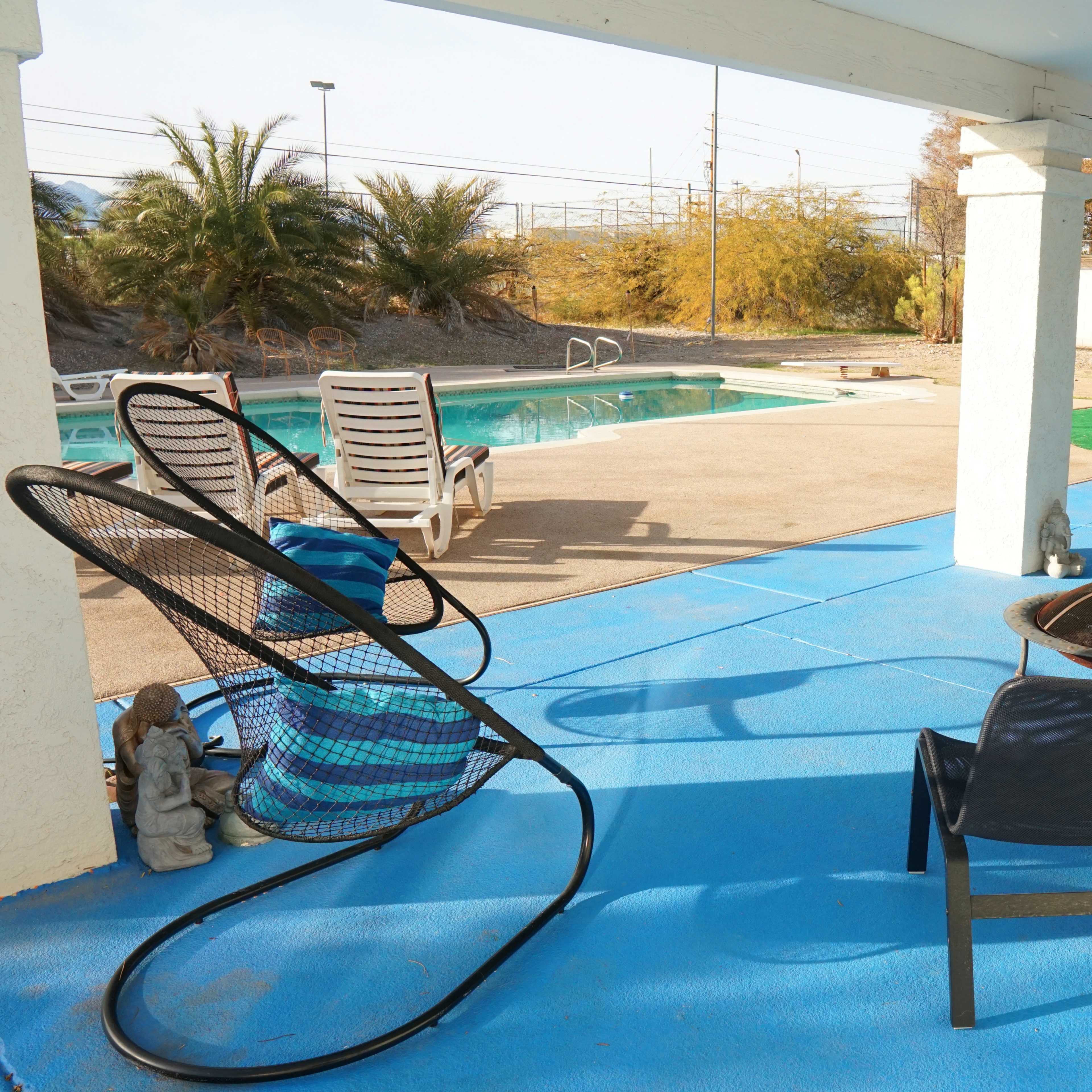A patio area with several chairs and a small table overlooking a swimming pool surrounded by palm trees.