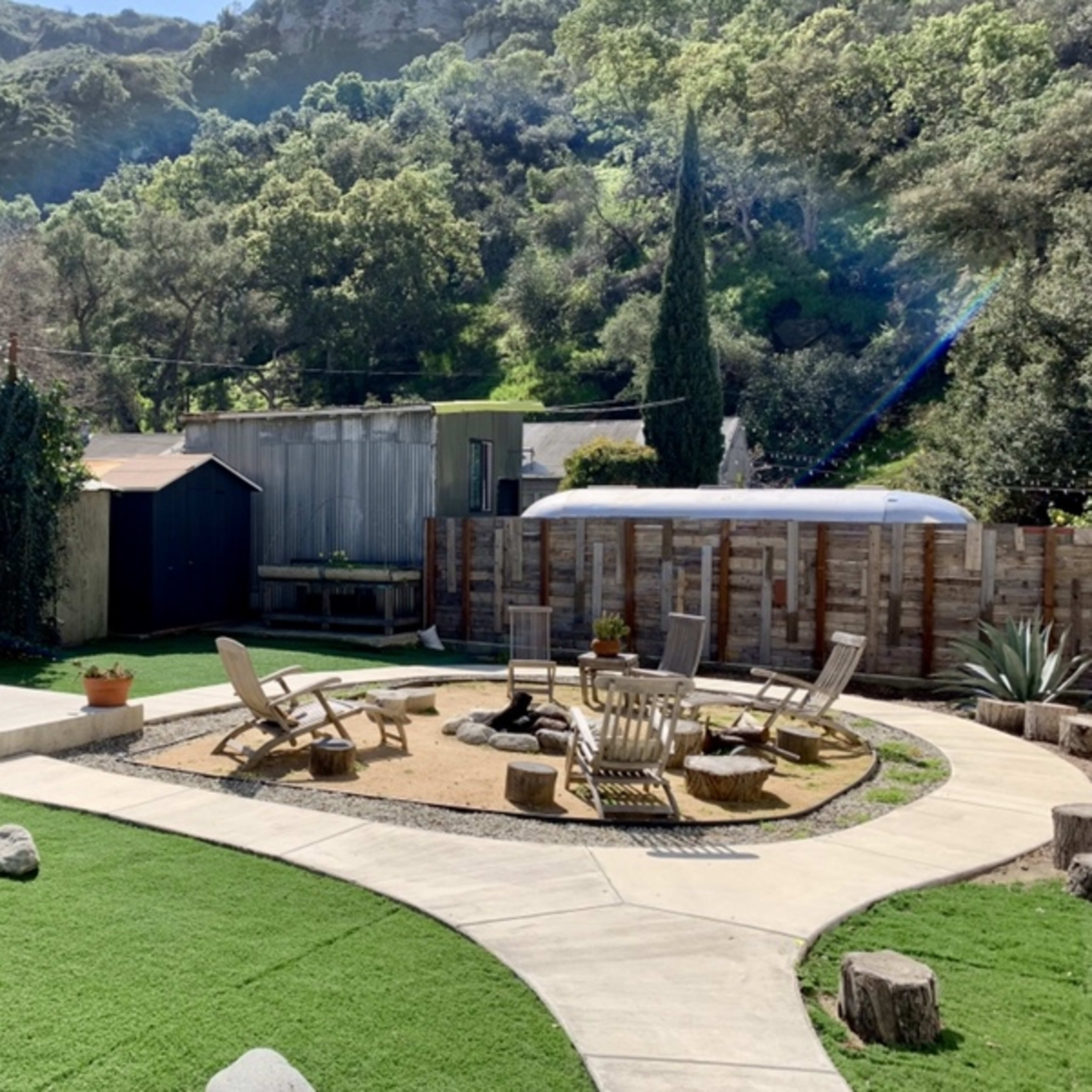 A landscaped outdoor area featuring seating around a fire pit, a grassy lawn, and a shed in a natural setting with hills in the background.