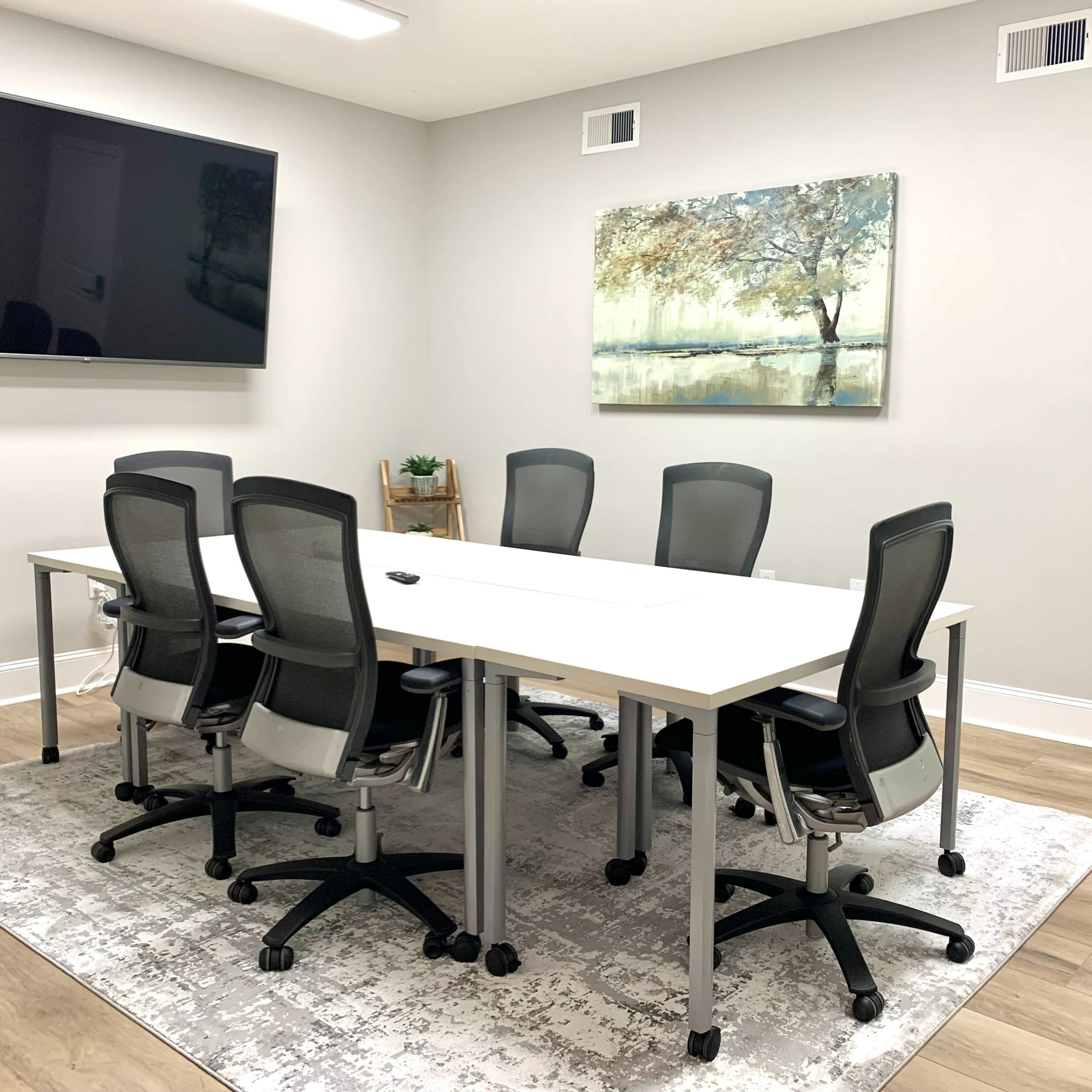 A modern conference room with a large table surrounded by six ergonomic chairs, a wall-mounted TV, and an artwork depicting a tree.