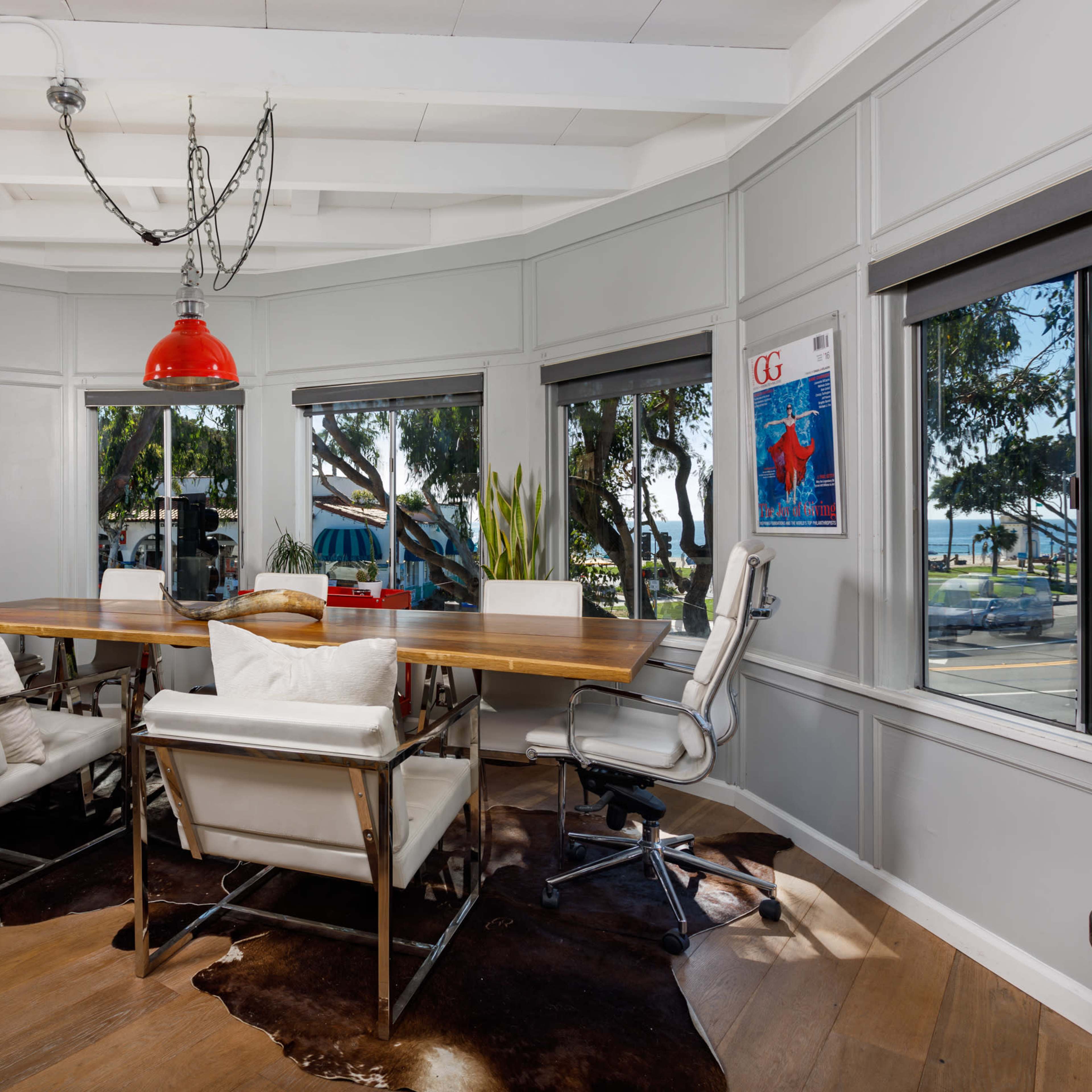 A modern conference room features a large wooden table surrounded by white chairs, with large windows offering a view of the outdoor scenery.
