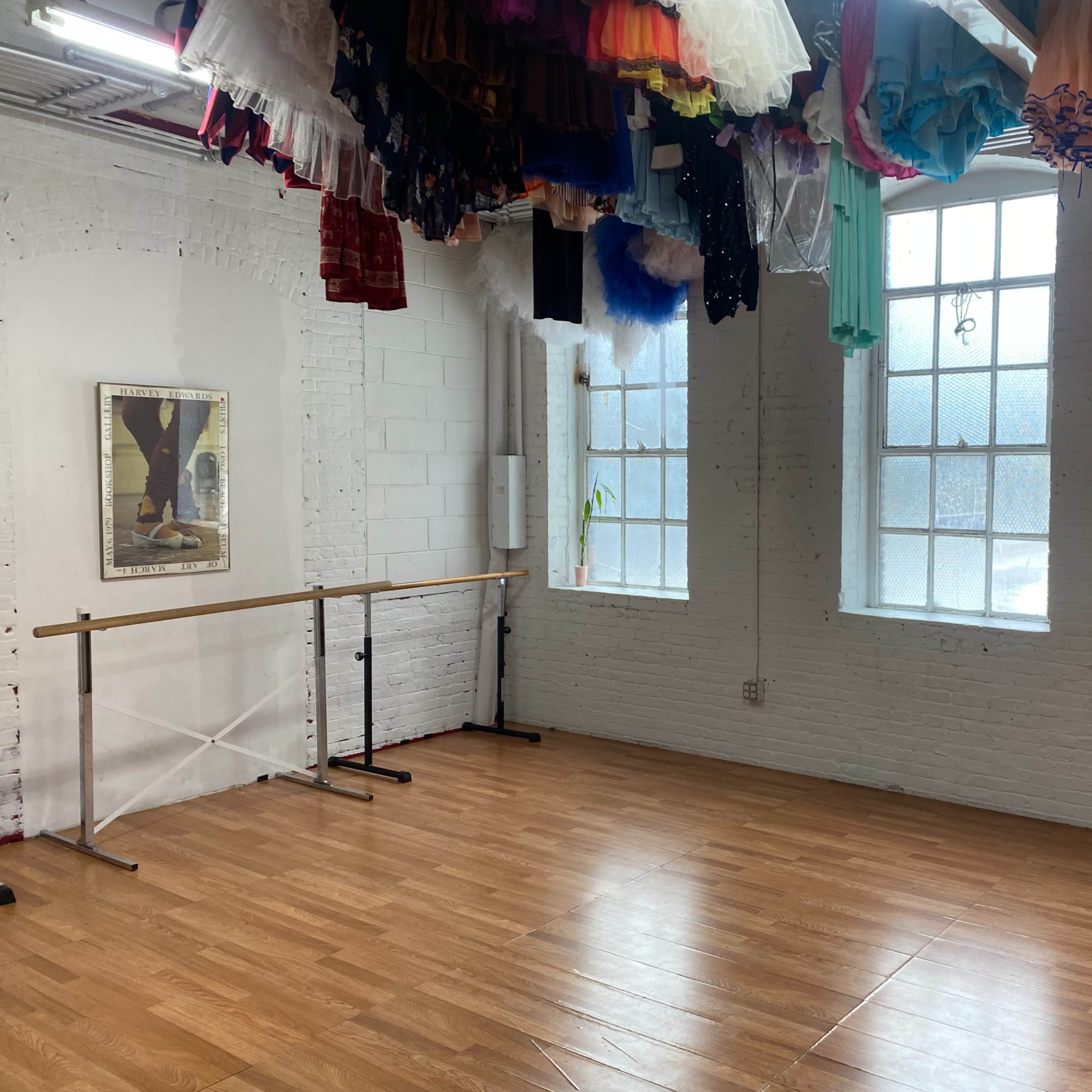 A brightly lit dance studio features wooden flooring, a ballet barre along one wall, and colorful costumes hanging from the ceiling.