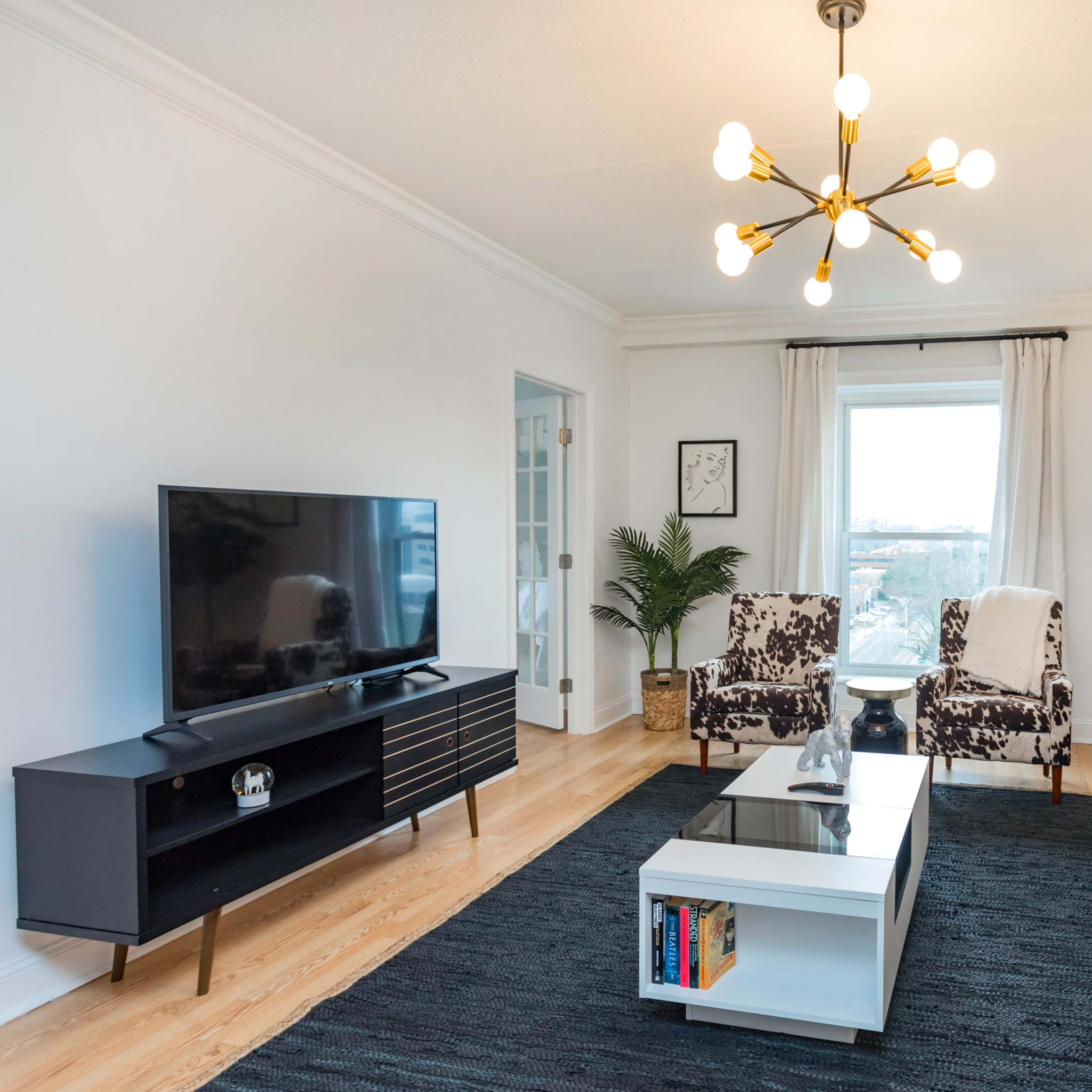 A modern living room features a gray sofa, a black television unit, a coffee table, and two patterned chairs, with a large window providing natural light.