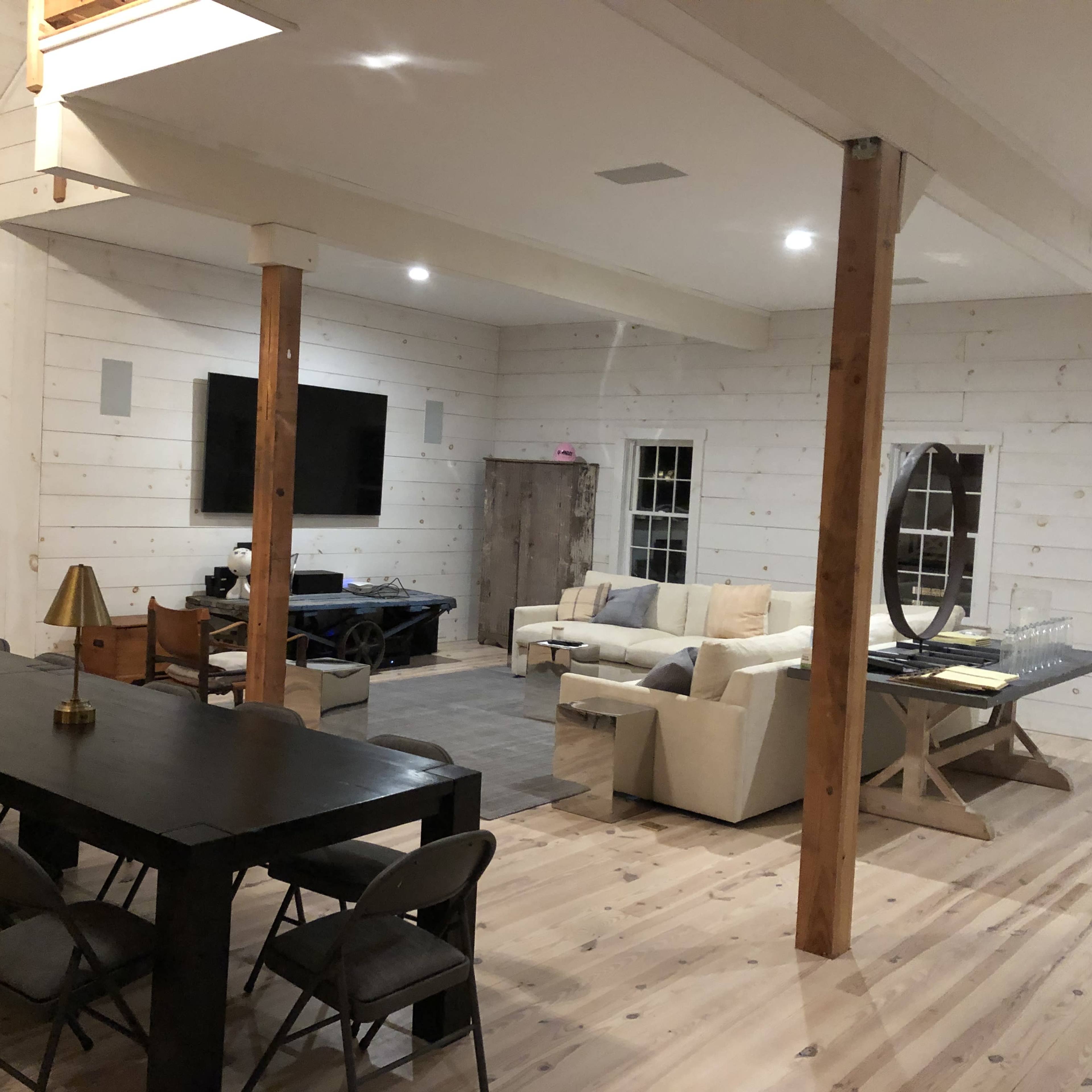 A spacious, modern interior with whitewashed wooden walls, a large dining table with chairs, a seating area with a light-colored couch, and a television mounted on the wall.