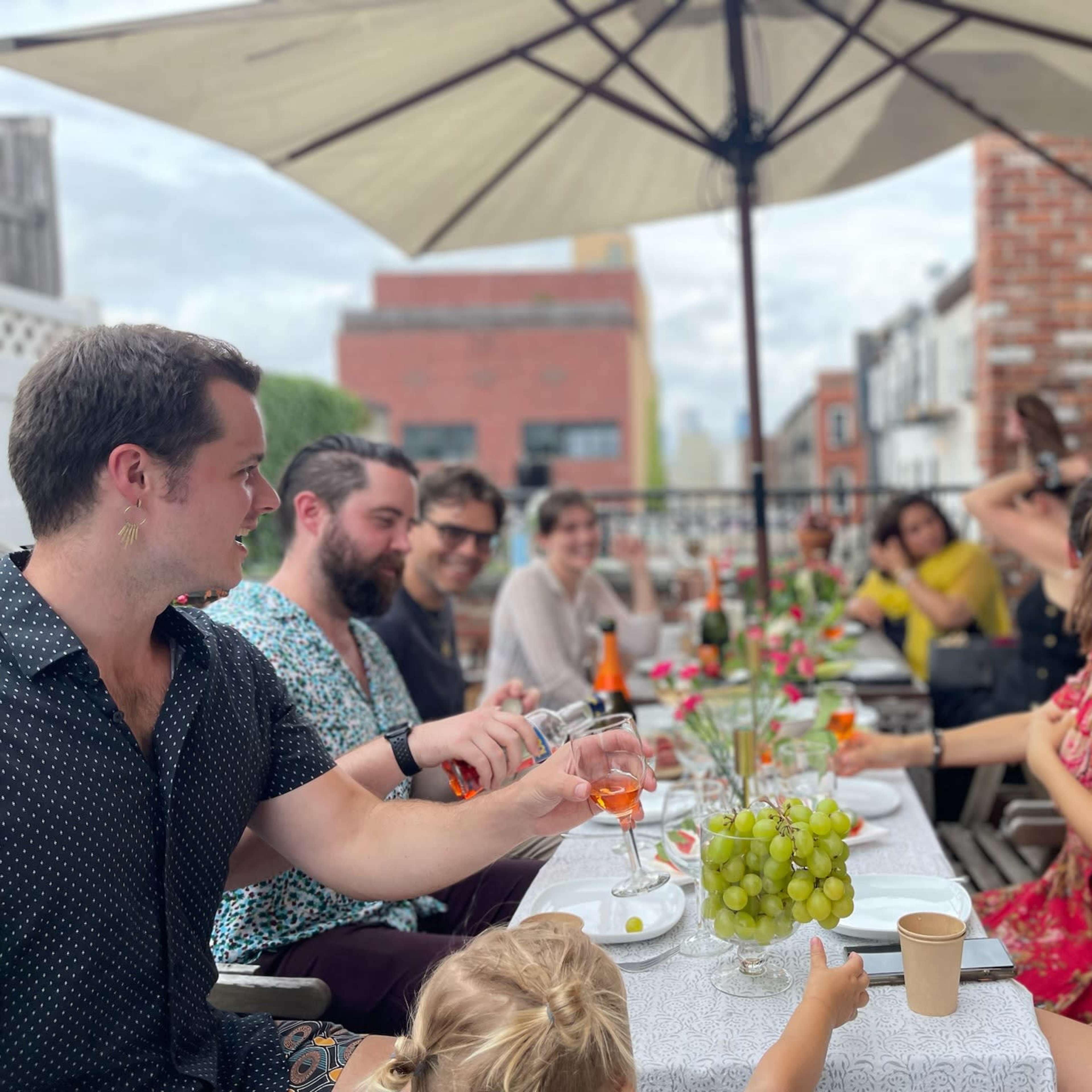 A group of people is seated around a long table adorned with food and drinks under an umbrella on a patio.