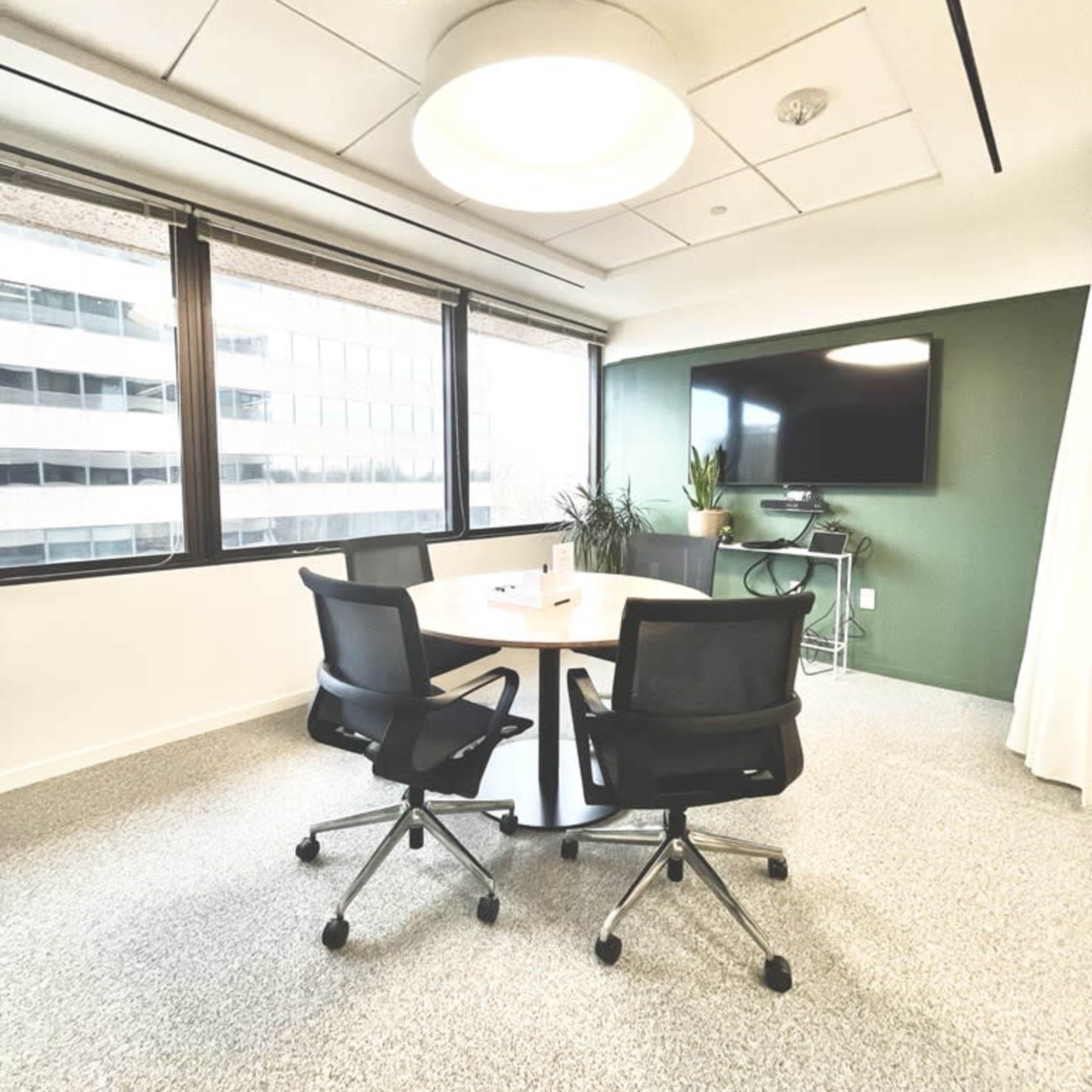 A modern conference room features a round table with three black chairs, large windows overlooking a cityscape, and a mounted TV against a green accent wall.