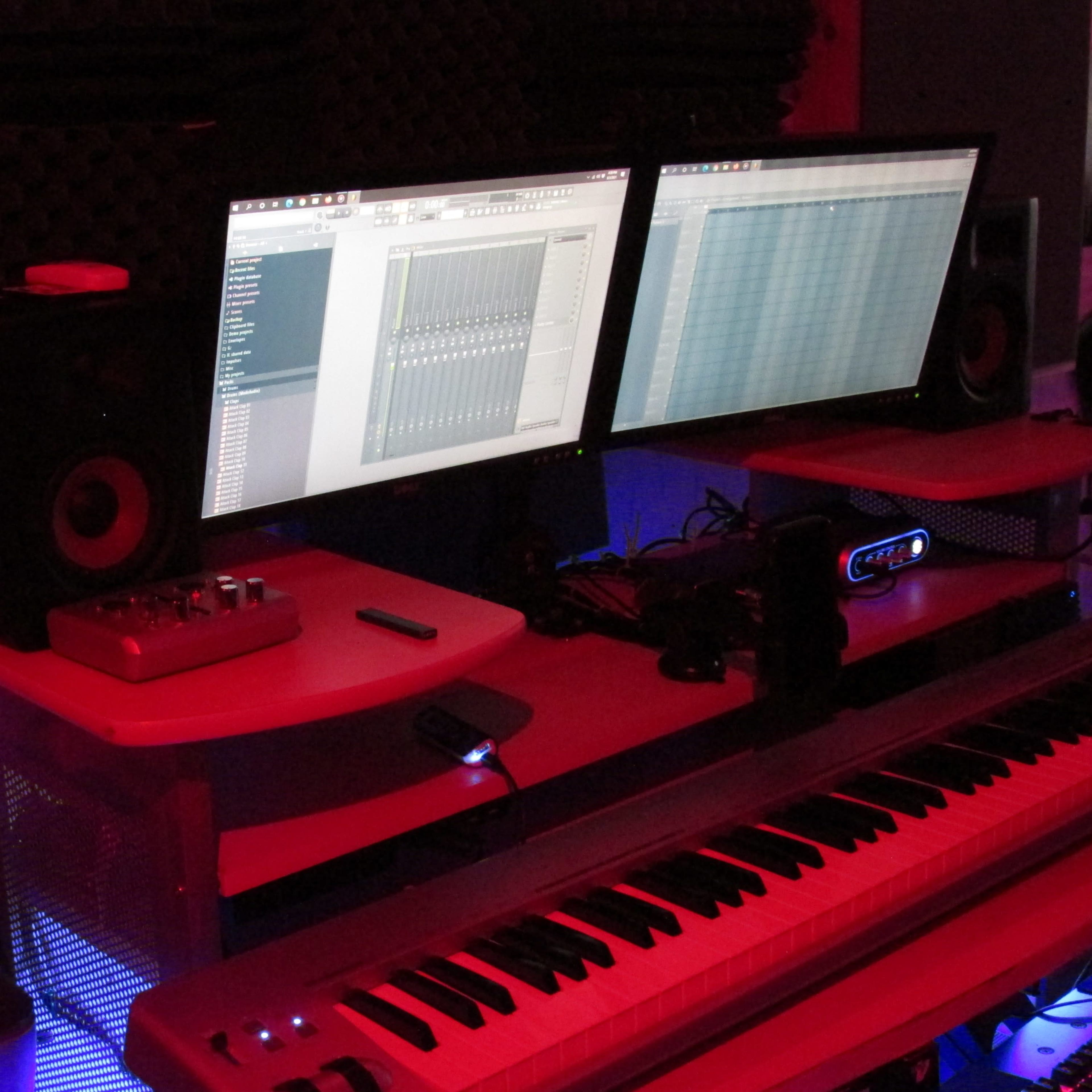 A music production setup features two monitors displaying digital audio software, a keyboard, and speakers in a dimly lit room.
