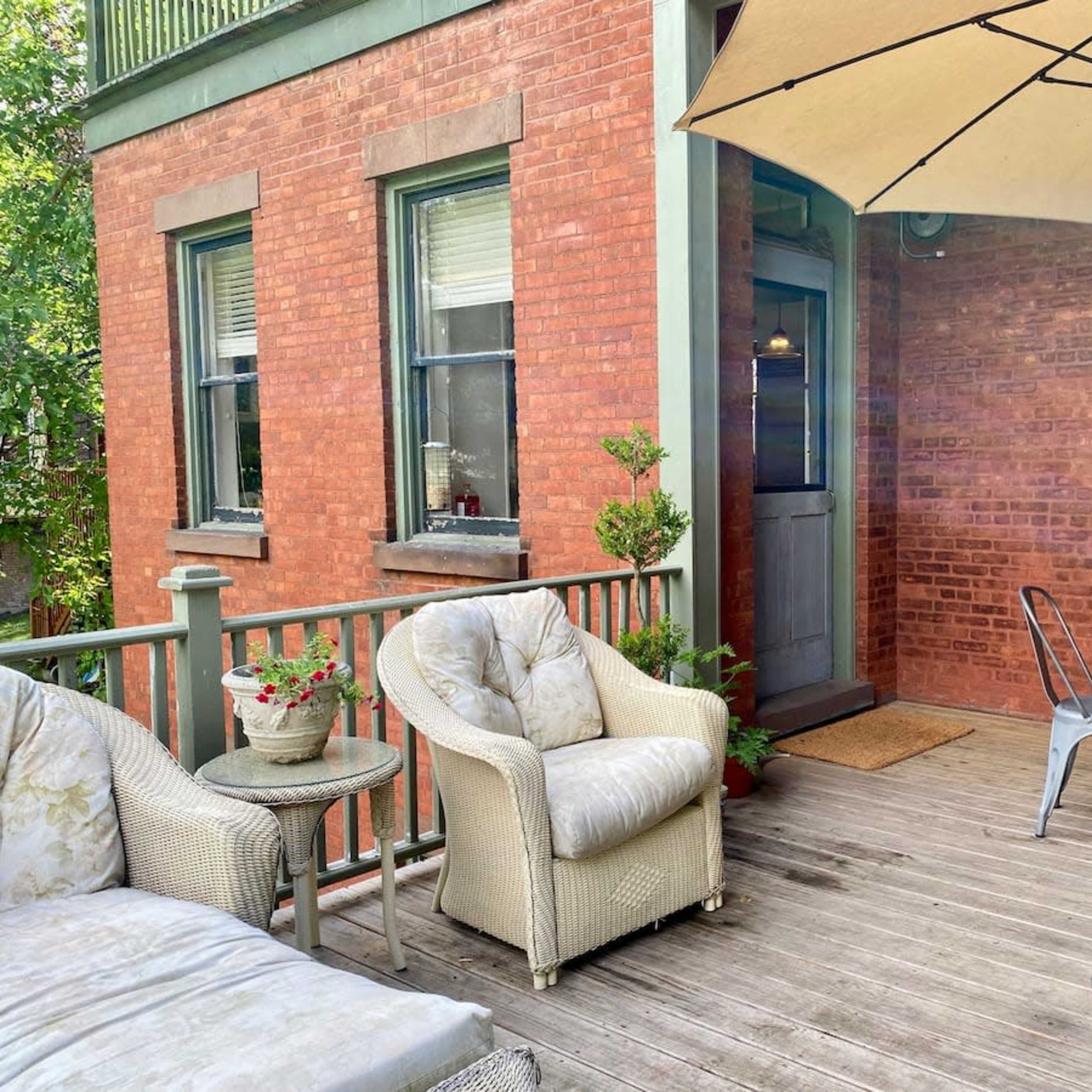 A wooden deck features a table with chairs, a large umbrella, and seating arrangements outside a brick building.