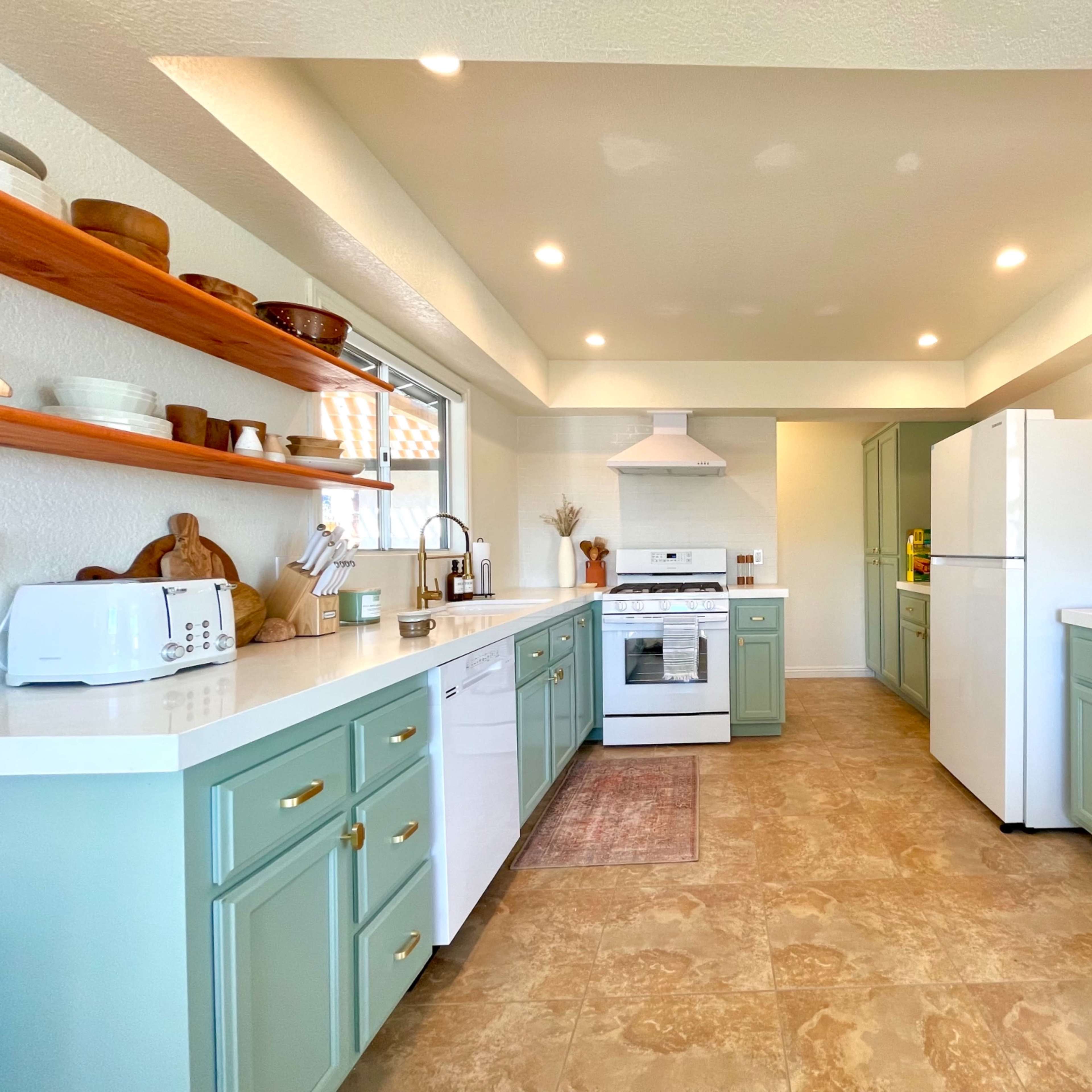 A modern kitchen with light green cabinets, white countertops, open shelves displaying kitchenware, and stainless steel appliances.