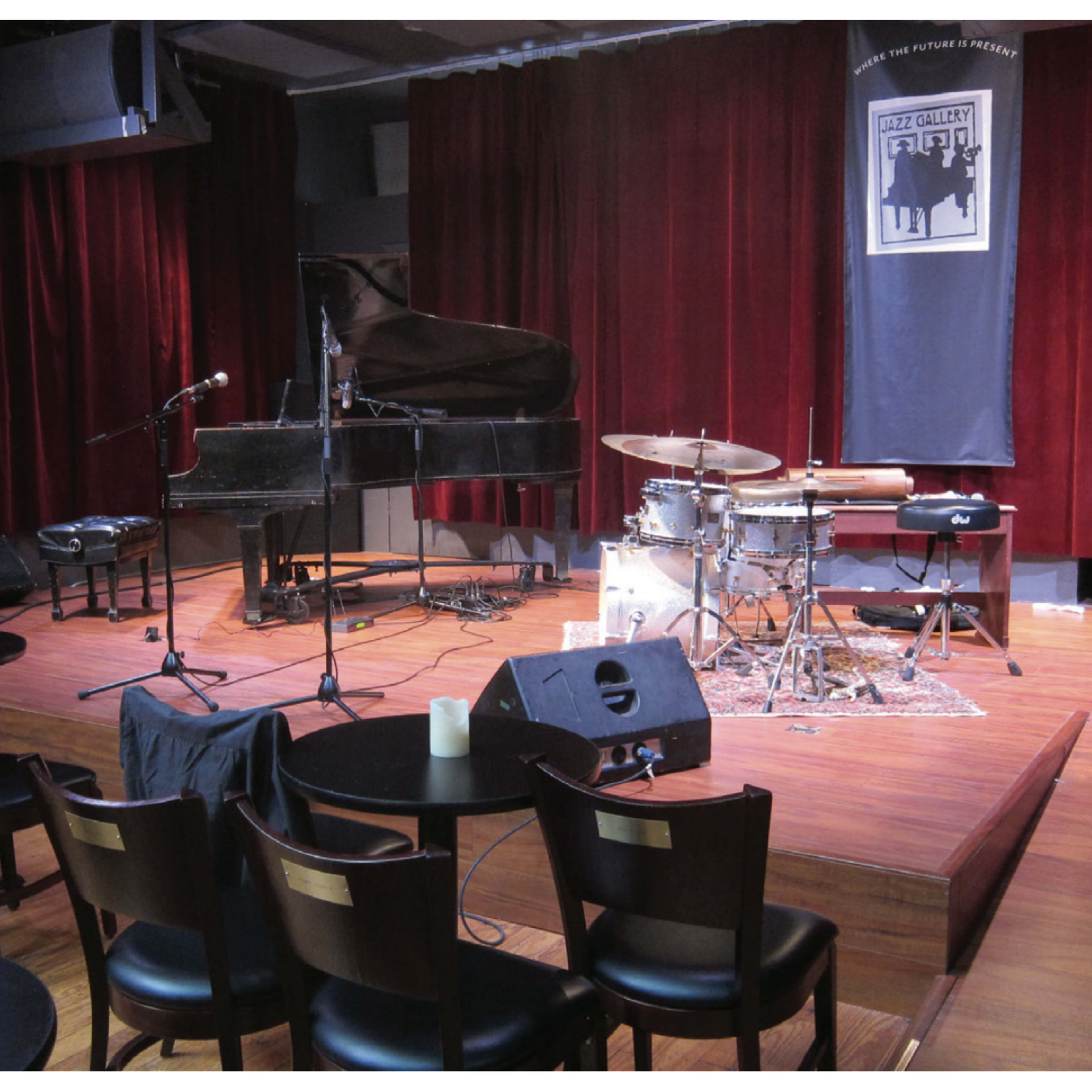 A small stage is set for a music performance, featuring a grand piano, a drum set, and microphones, surrounded by chairs and dim candlelight.