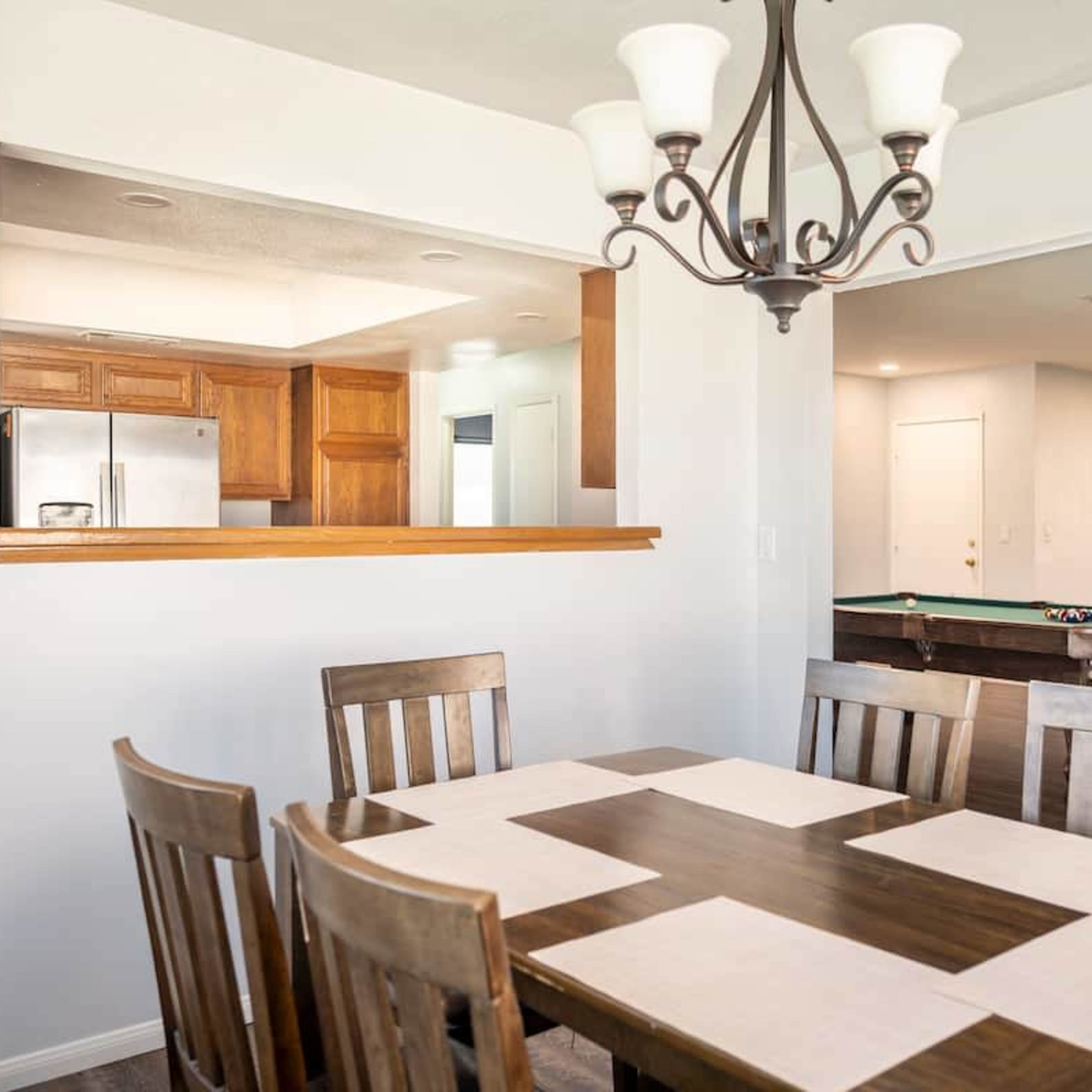 A dining area features a wooden table with placemats, surrounded by chairs, and a view into an adjacent kitchen with wooden cabinets and an open doorway leading to a room with a pool table.