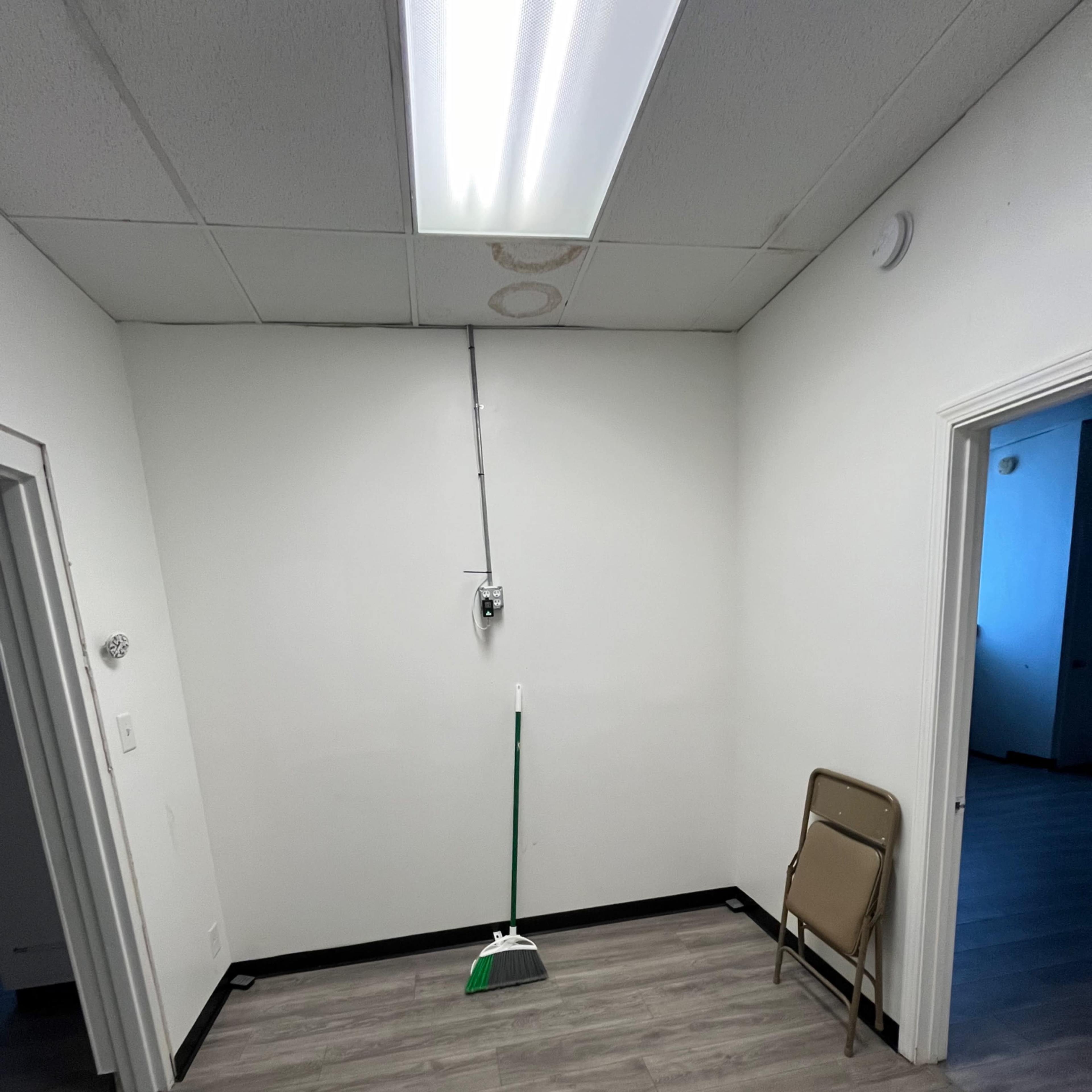 A small room with a broom leaning against the wall, a folding chair in the corner, and two doorways leading to other spaces.
