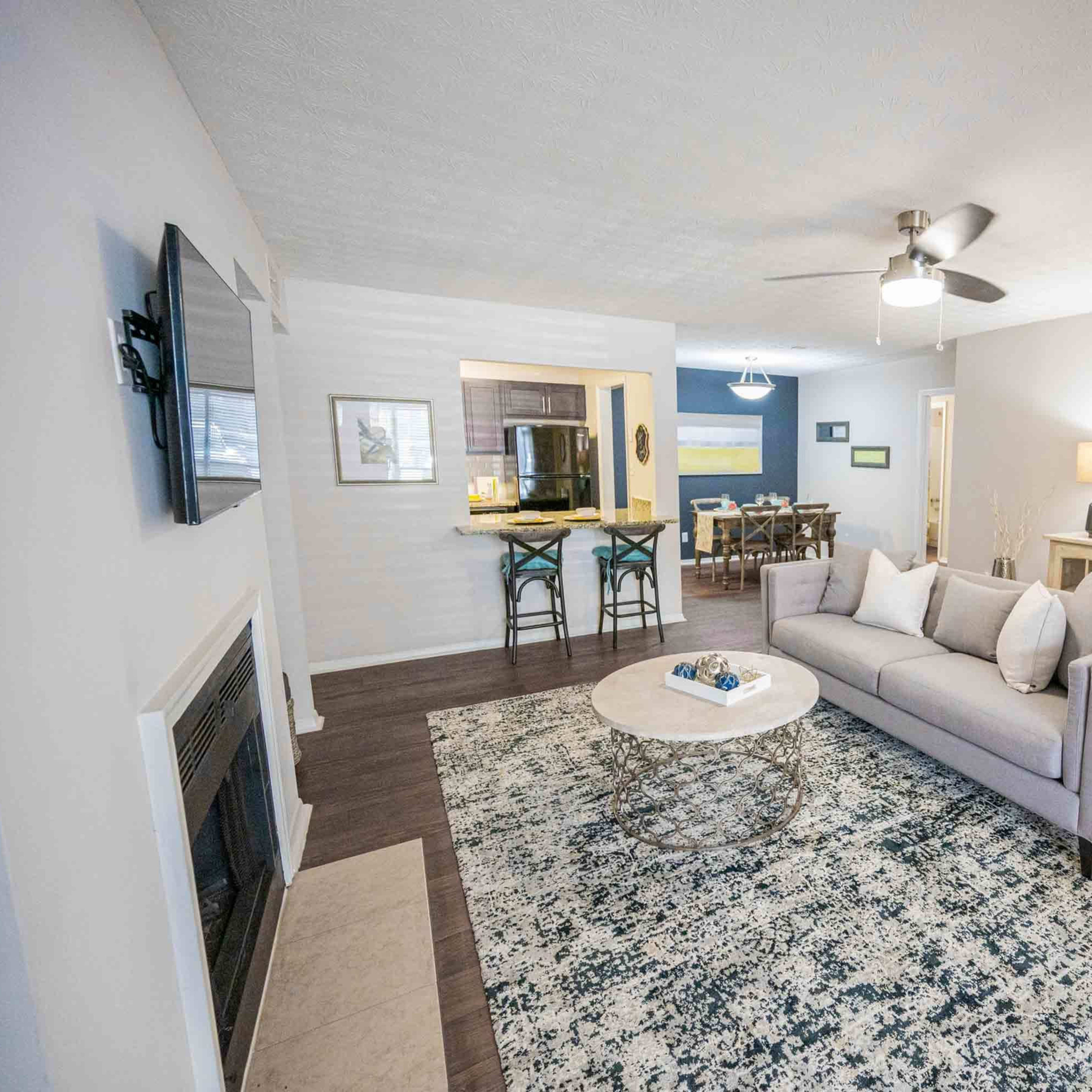 The living room features a gray sofa facing a circular coffee table on a patterned rug, with a television mounted on the wall and an open kitchen visible in the background.