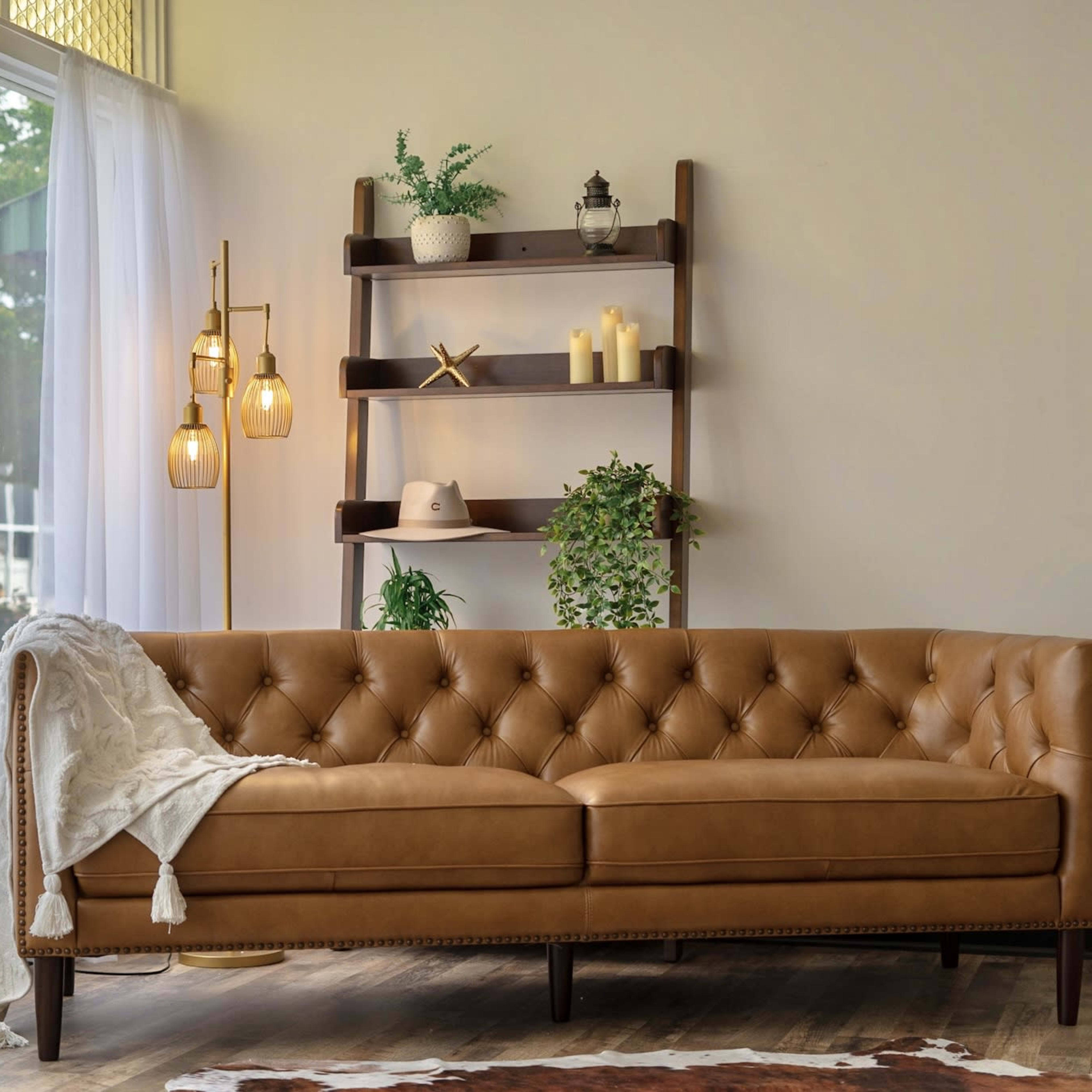 A tan tufted sofa is positioned in front of a wooden shelving unit with decorative plants and candles, beside sheer white curtains.