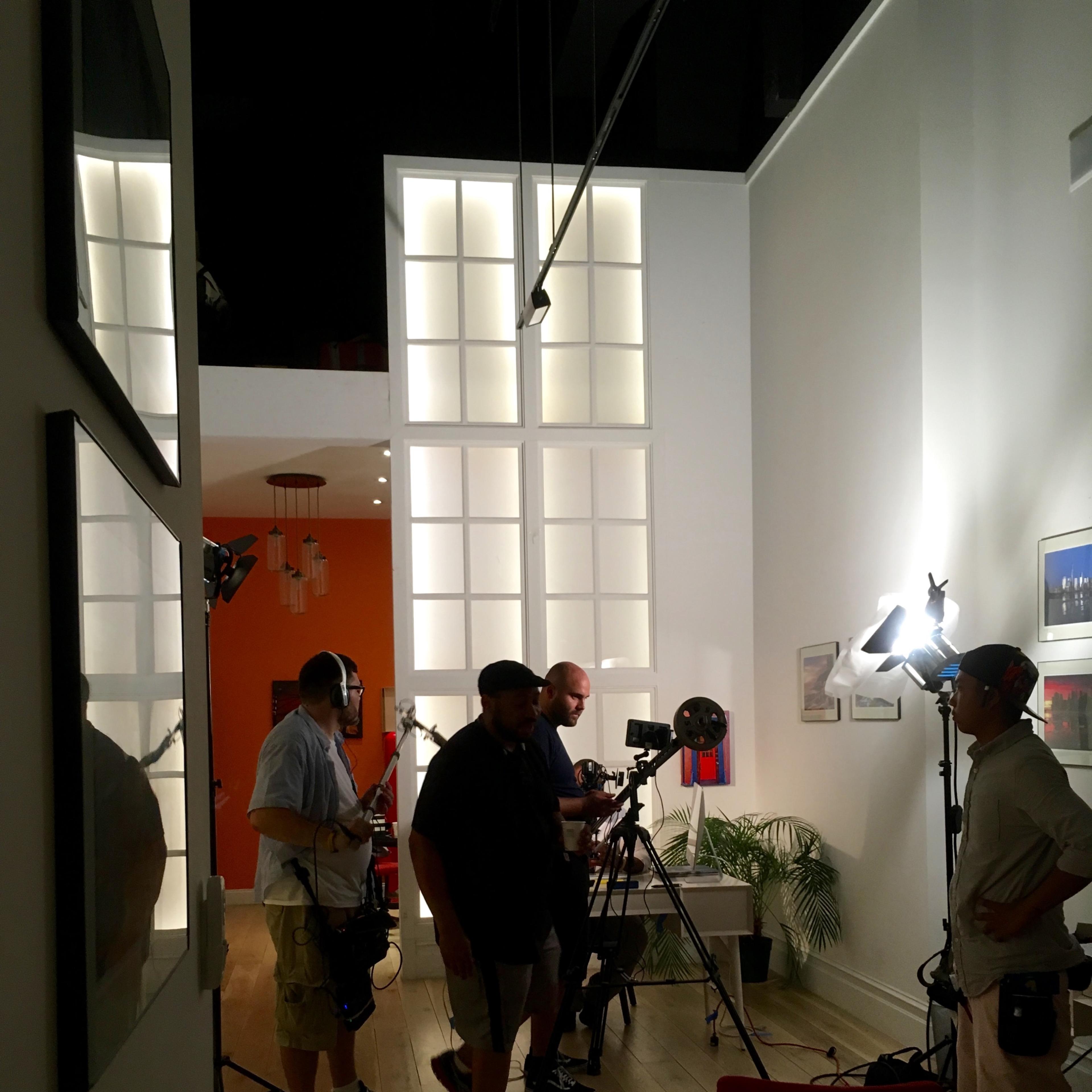 A group of four individuals sets up filming equipment in a well-lit, modern room with large windows and minimalistic decor.