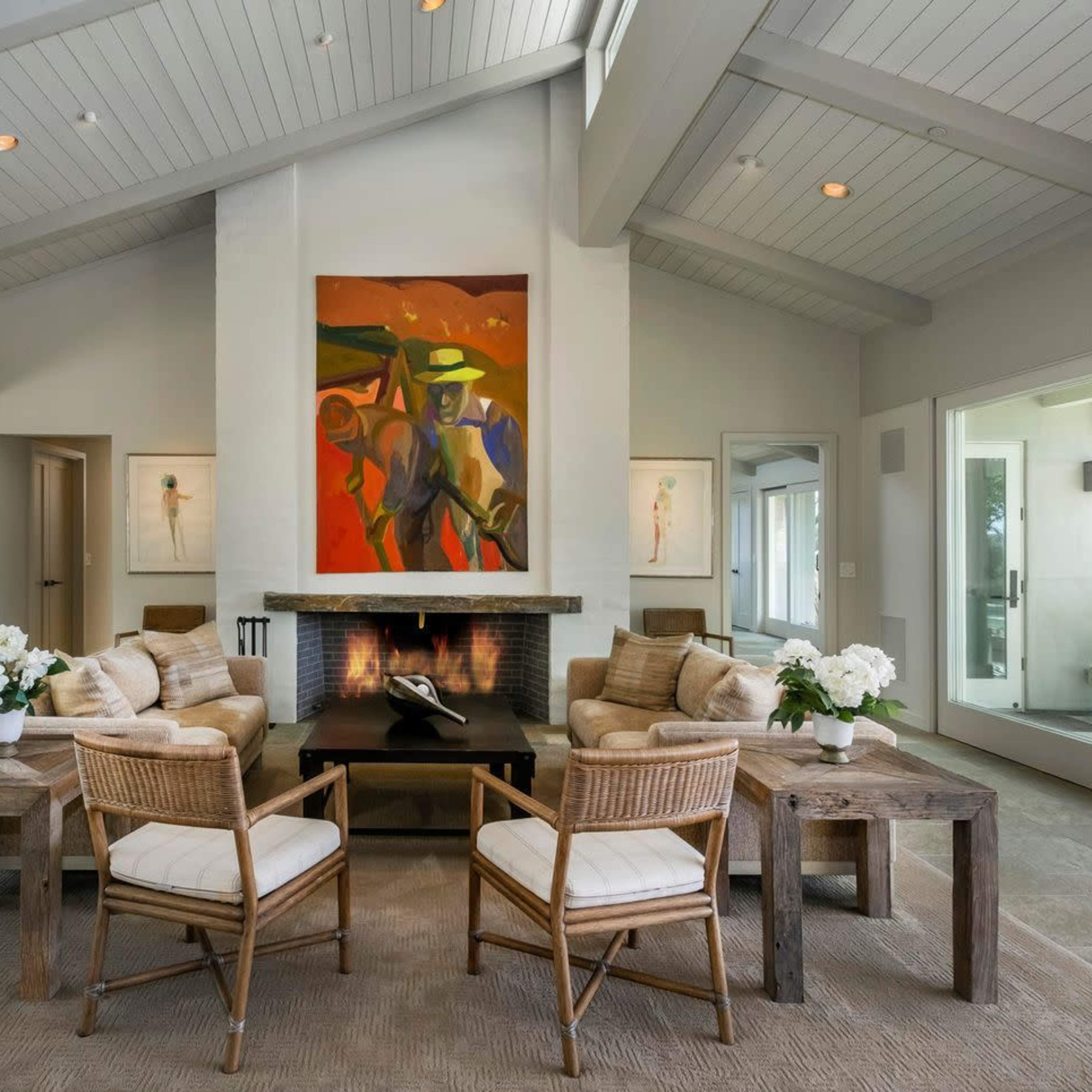 A modern living room featuring two armchairs and a coffee table arranged around a fireplace, with a large abstract painting on the wall and large windows allowing natural light.