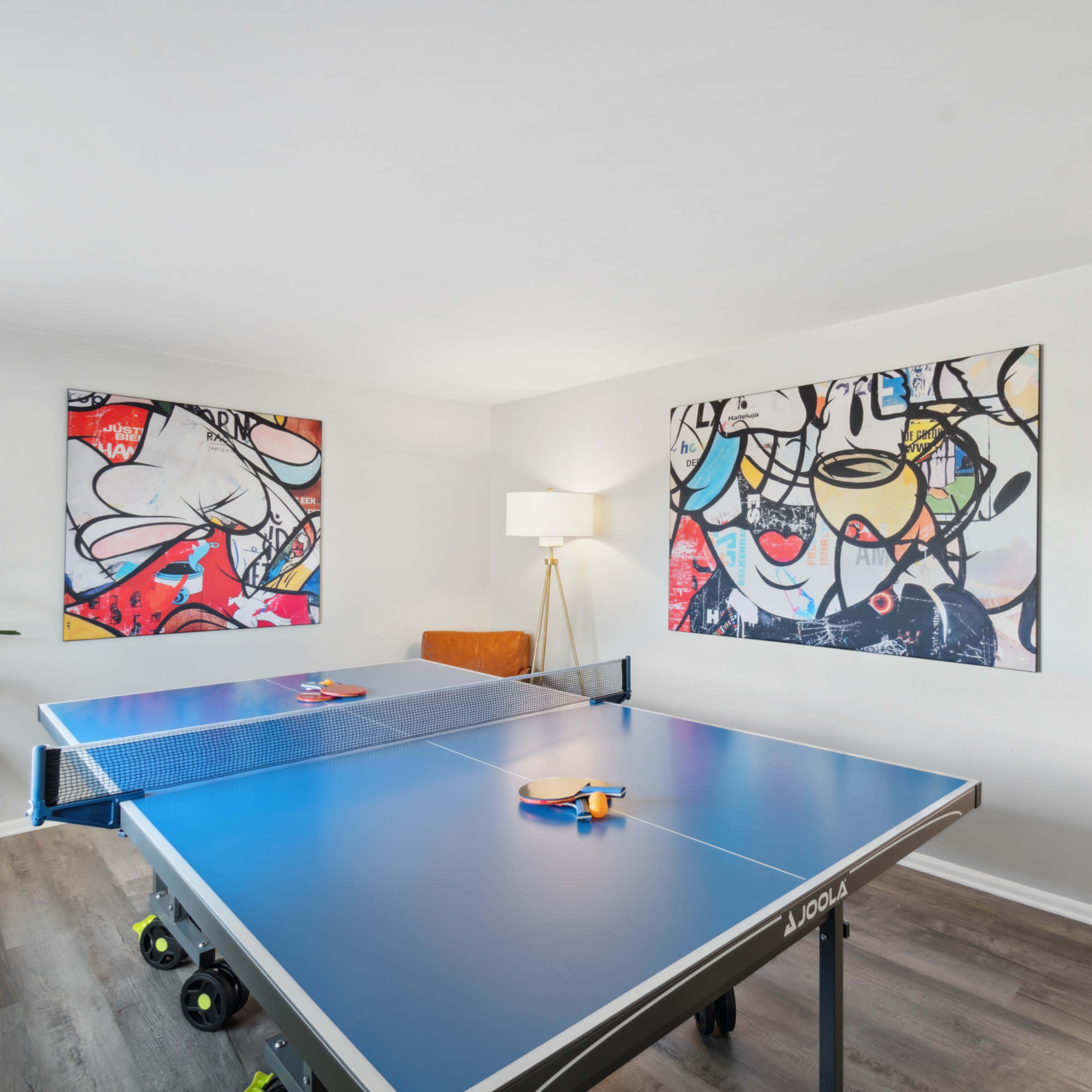 A bright room with a blue ping pong table in the center, surrounded by abstract art on the walls and a lamp beside the table.