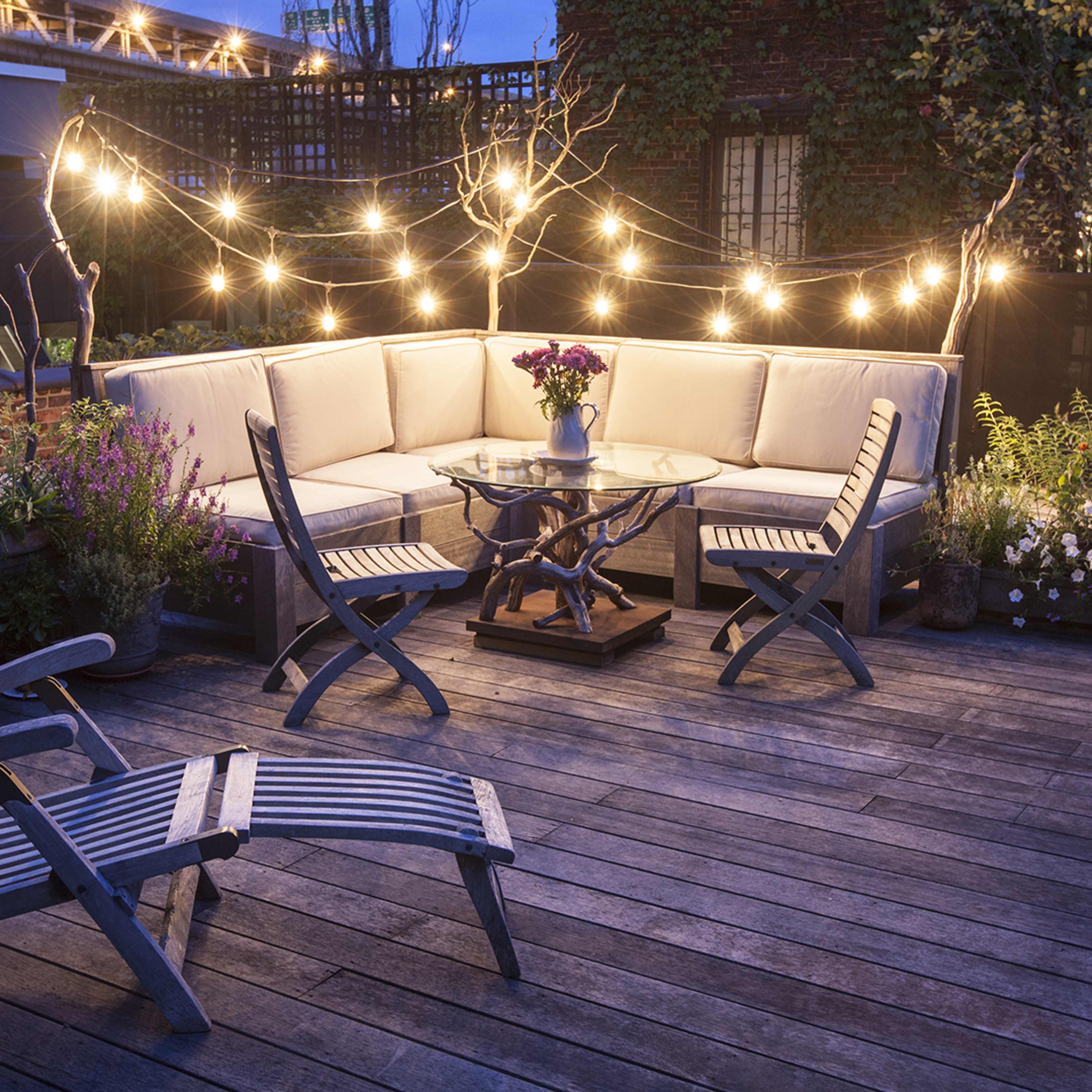 A cozy rooftop seating area features a sectional sofa, a glass table, and string lights, surrounded by potted plants and a wooden deck.