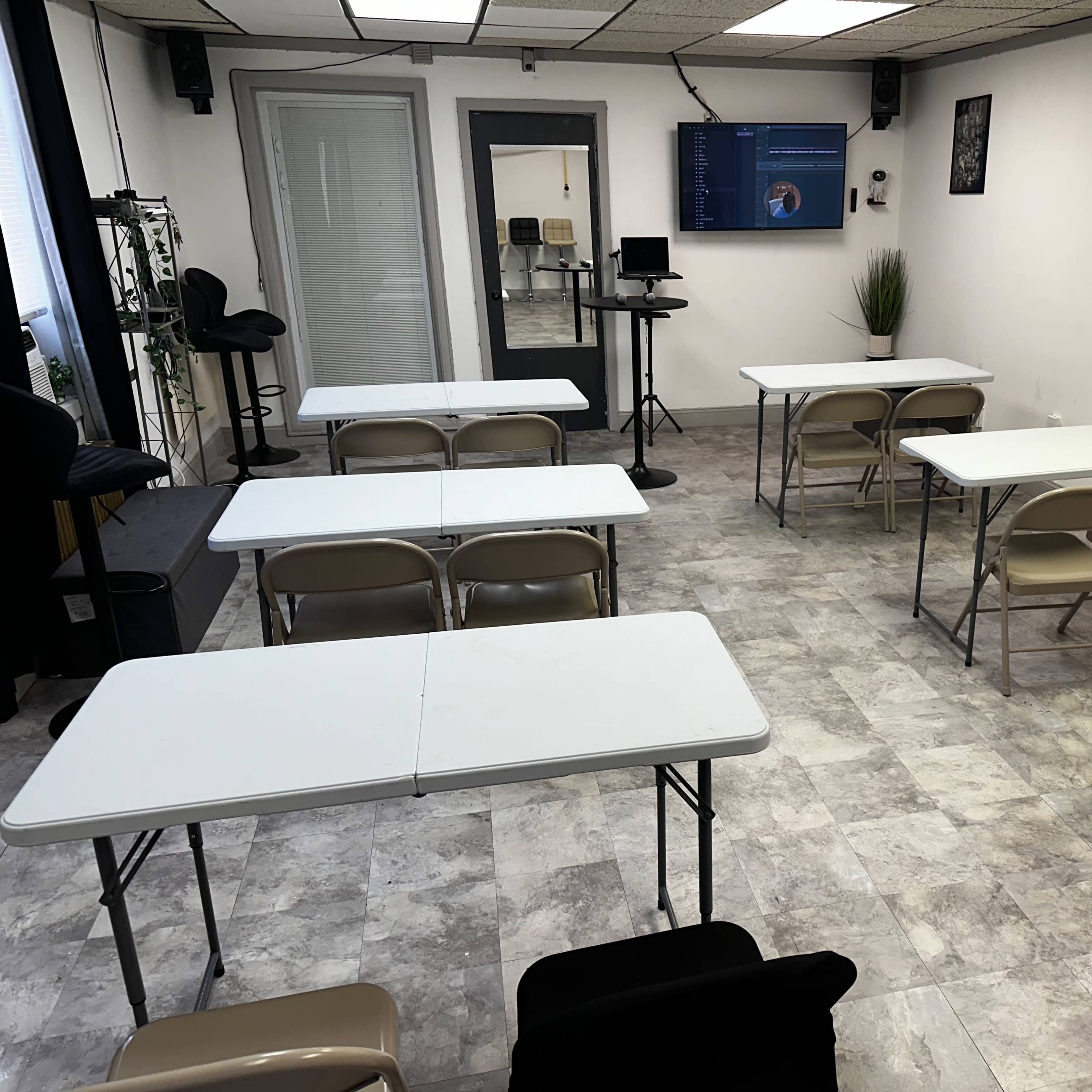 A classroom features six white tables arranged with folding chairs, and a large screen displaying content in the background.