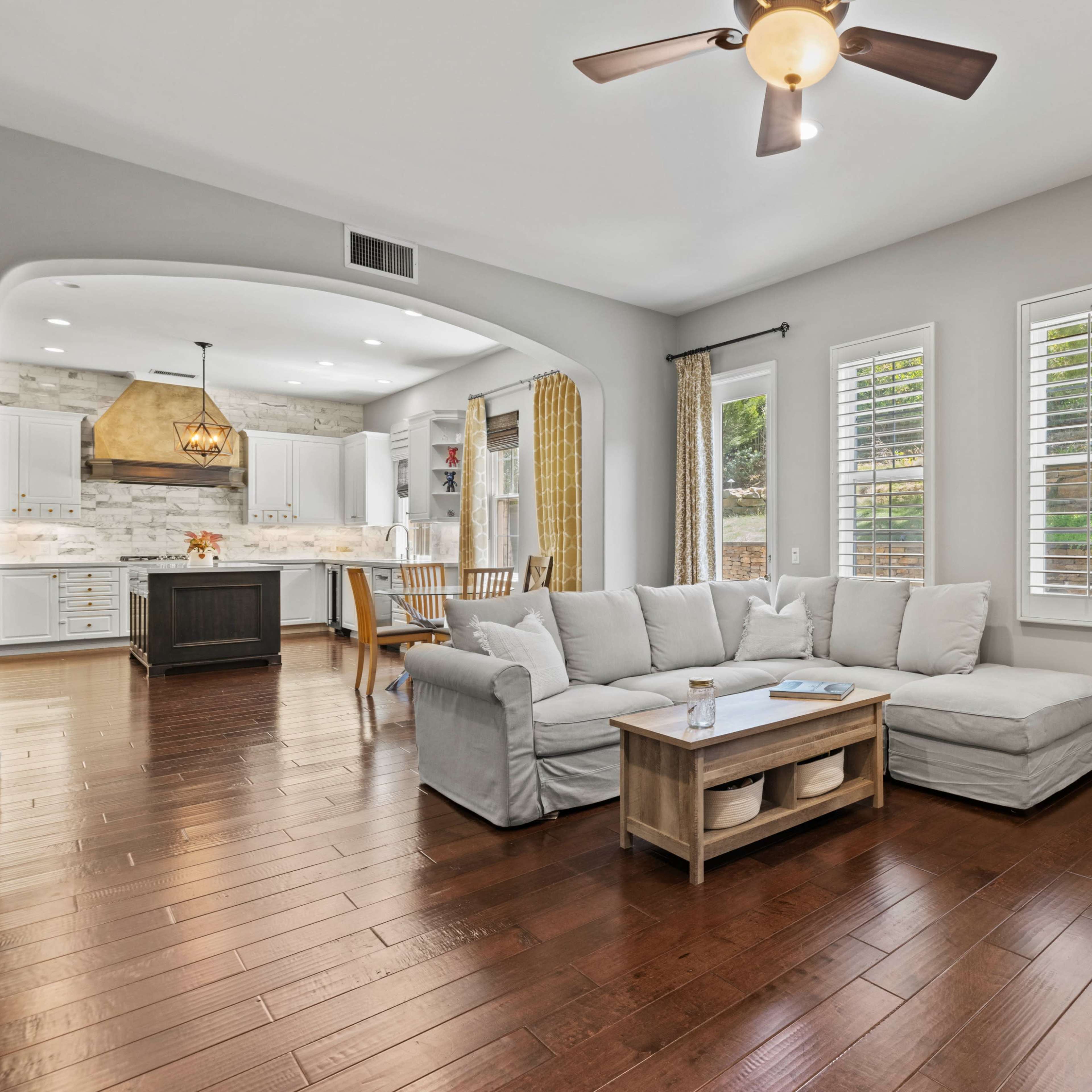 A living room with a light-colored sectional sofa, wooden flooring, and large windows letting in natural light, leading into a kitchen area with a pendant light.