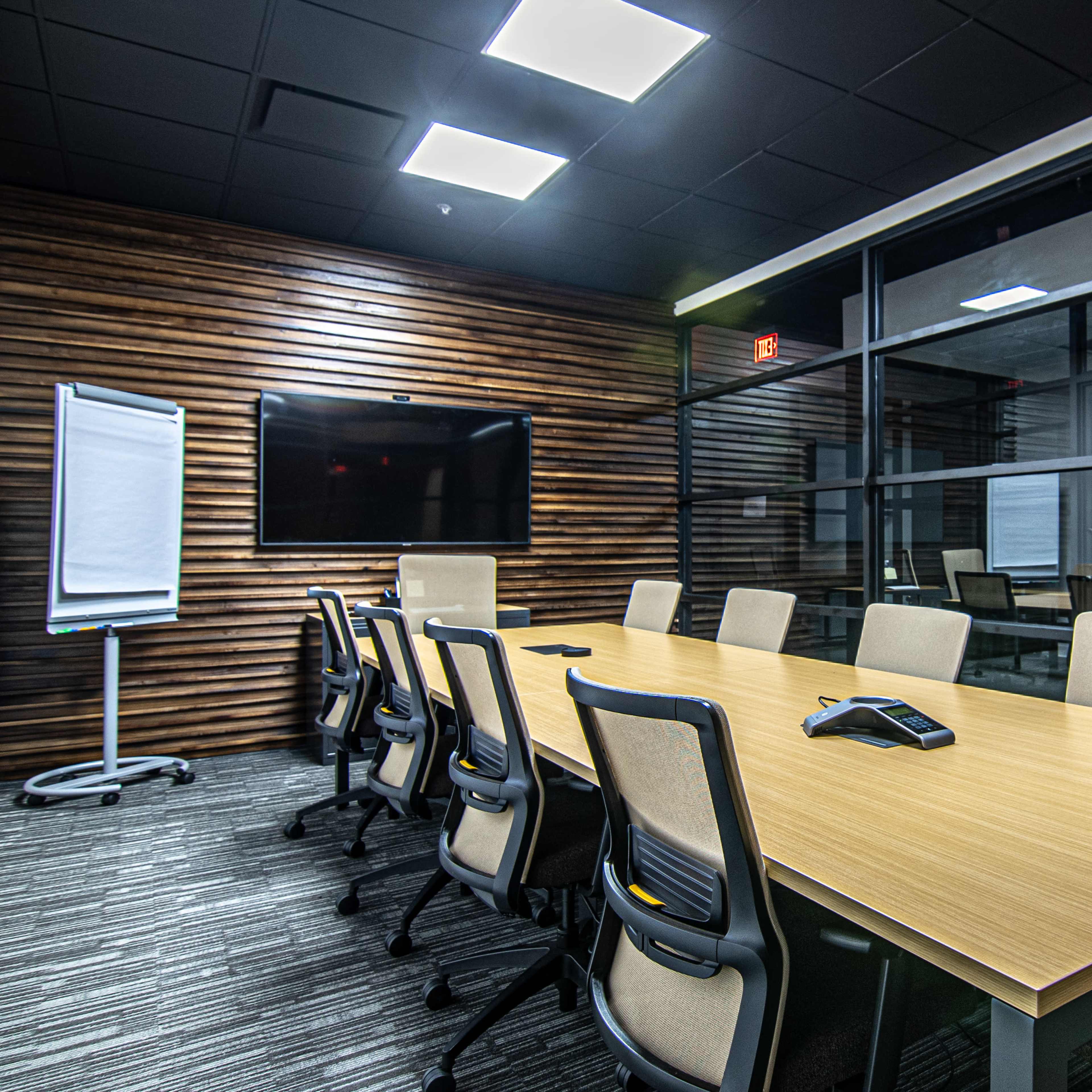 A large conference room features a rectangular wooden table surrounded by ergonomic chairs, with a wall-mounted screen and a glass partition.