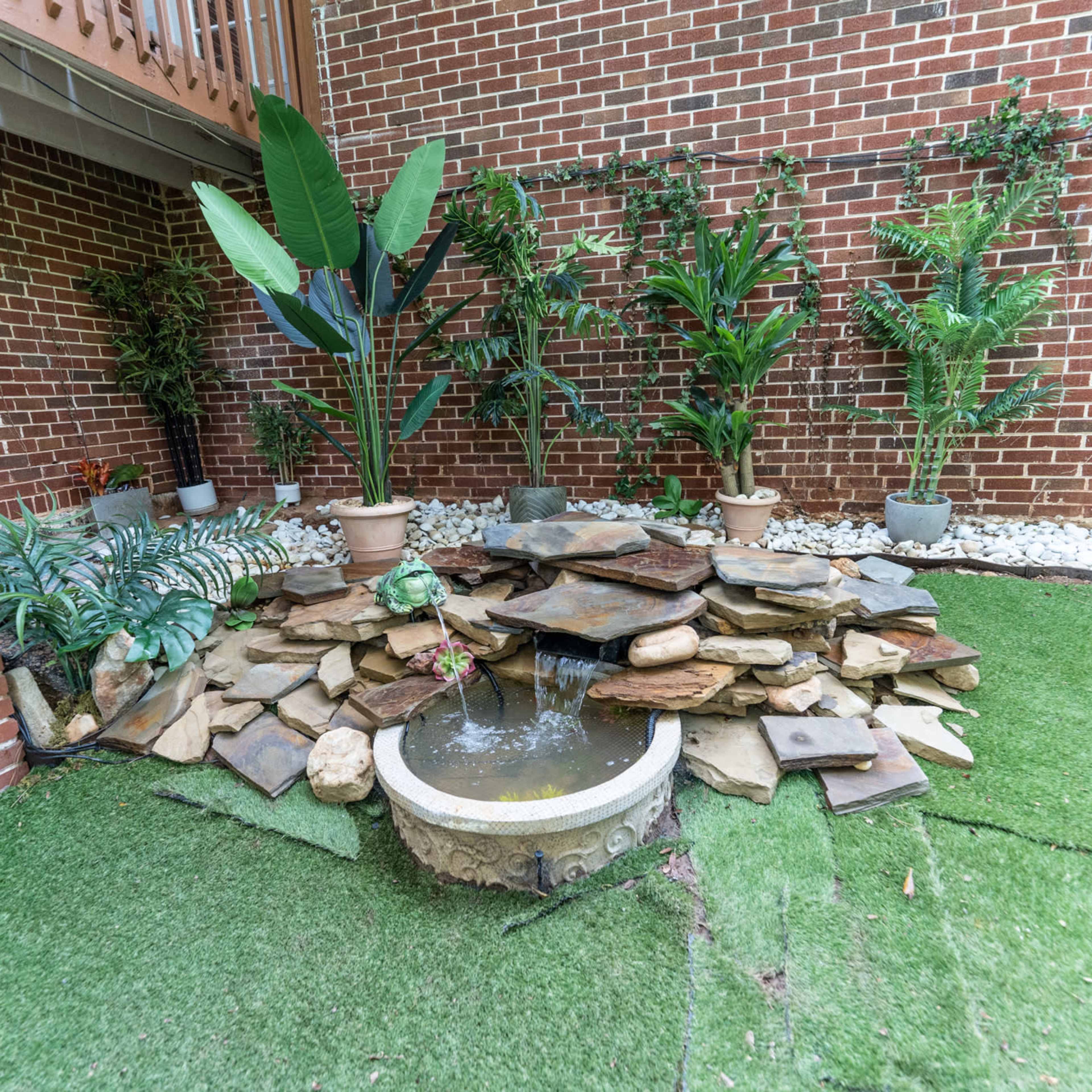 A landscaped corner with a stone fountain surrounded by potted plants and artificial grass against a brick wall.