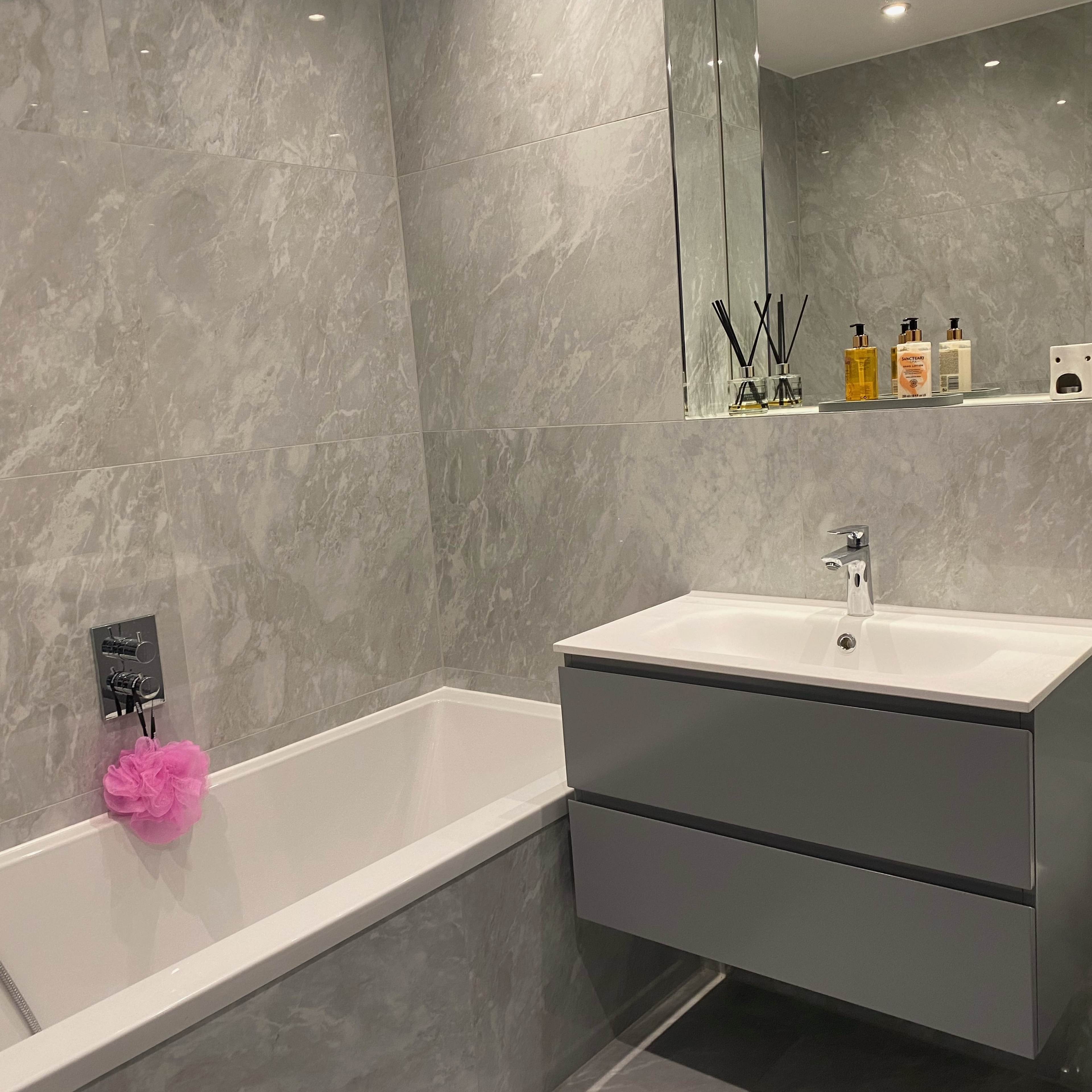 A modern bathroom with gray marble walls, a bathtub, a sink with a cabinet below, and various toiletries on the counter.