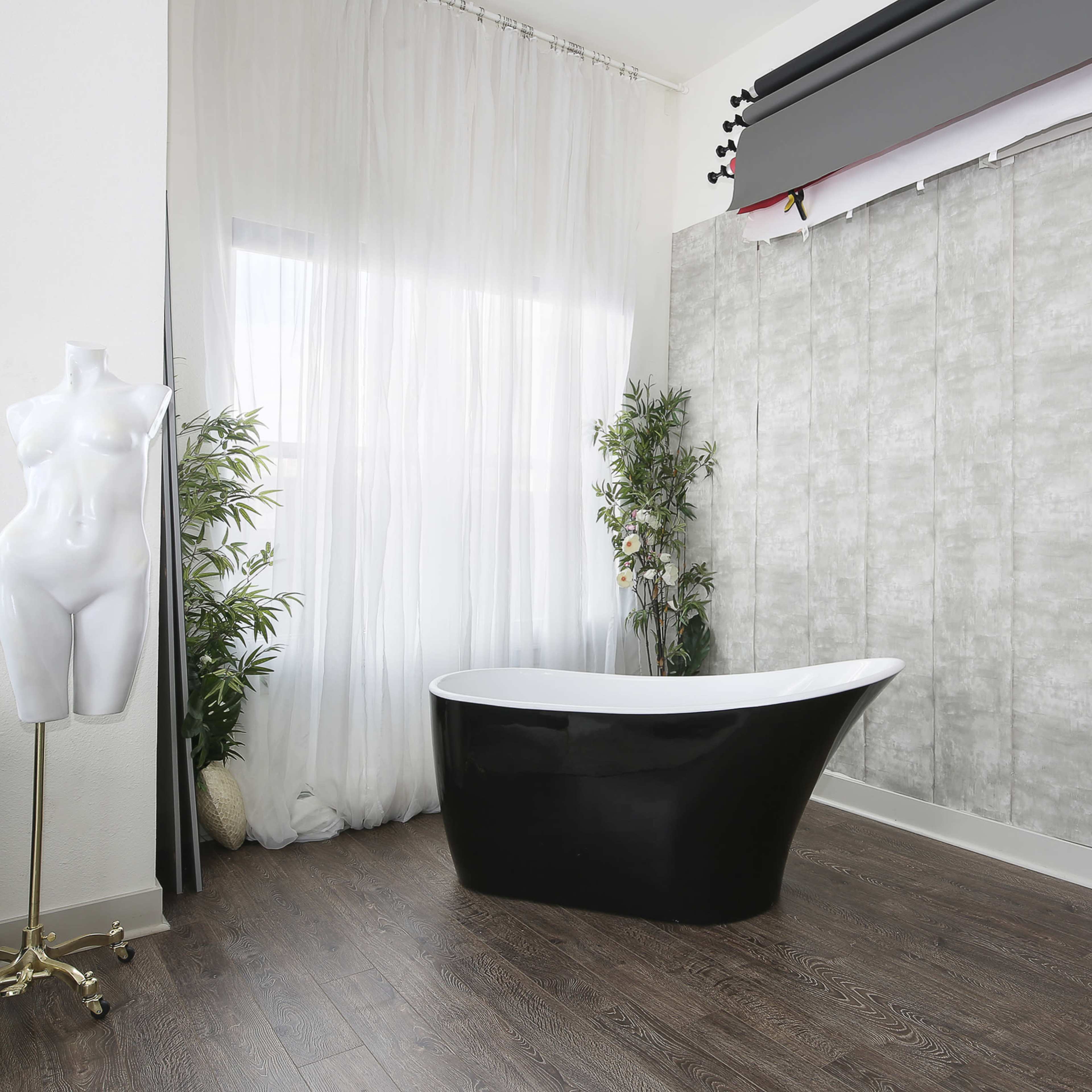 A modern bathroom with a black freestanding bathtub, two white mannequin forms, and large windows draped with sheer curtains.