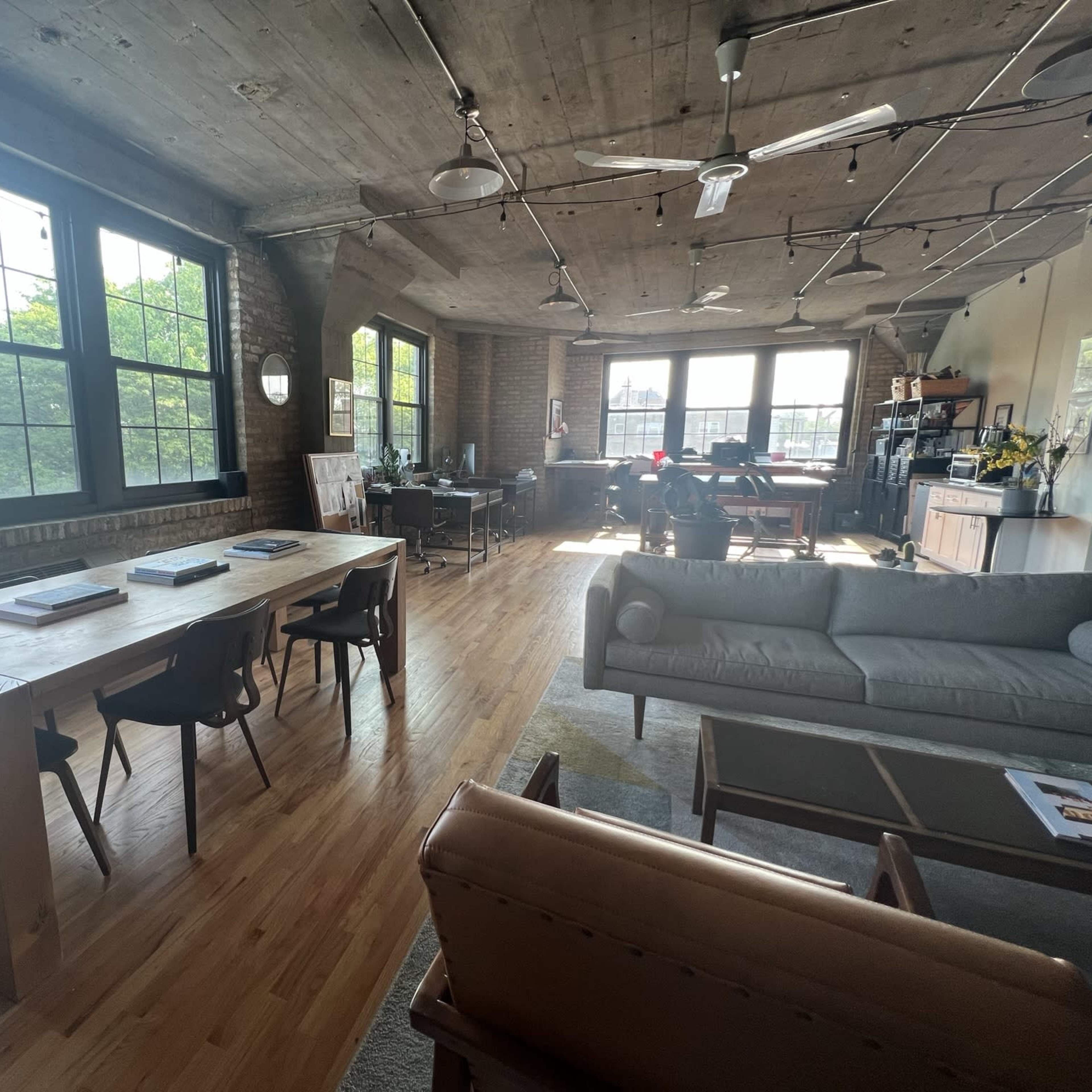 An open-concept loft space featuring a wooden dining table, a sofa, and a workspace with large windows allowing natural light to fill the room.