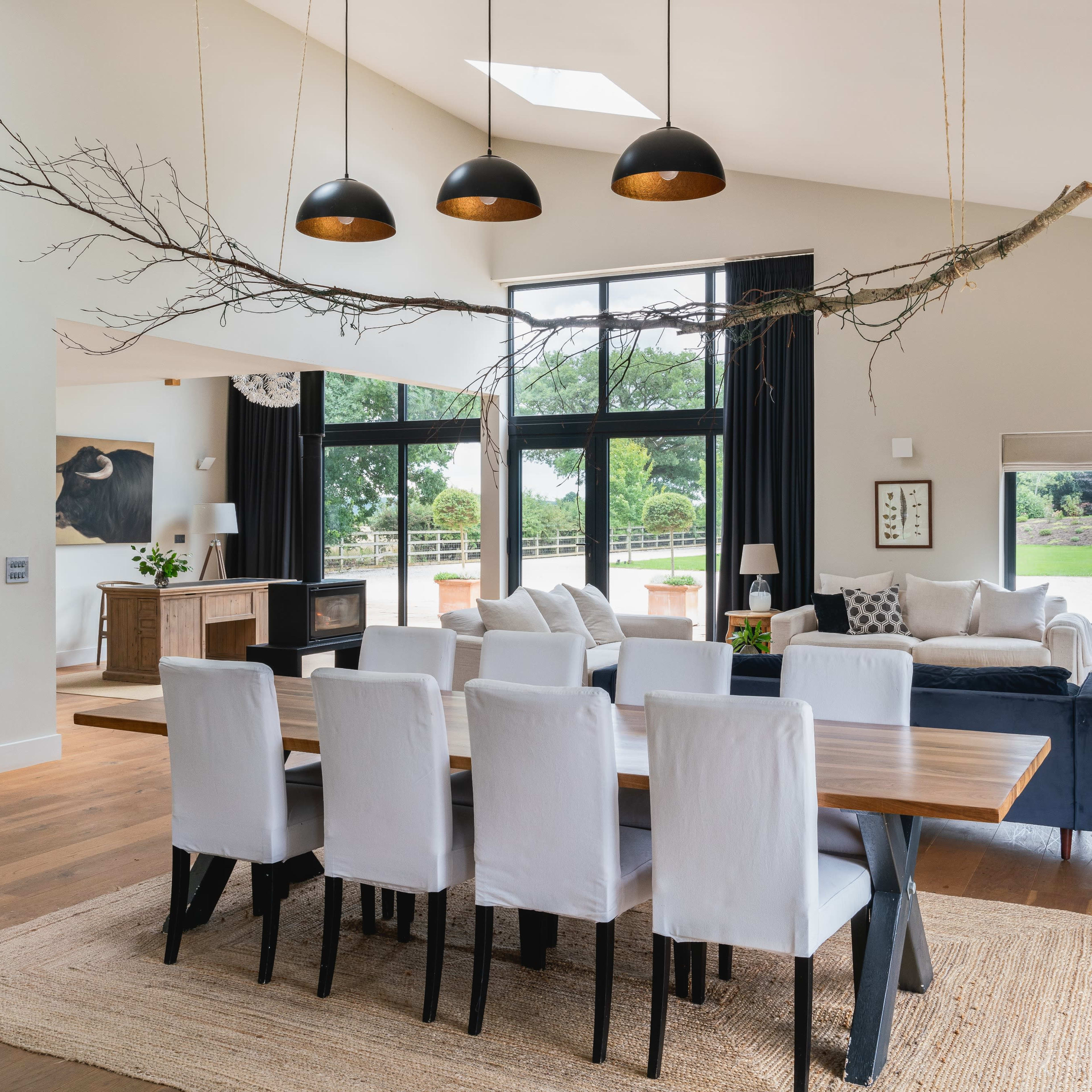 A spacious, modern dining area features a long wooden table surrounded by white chairs, with a branch light fixture overhead and large windows allowing natural light to fill the room.