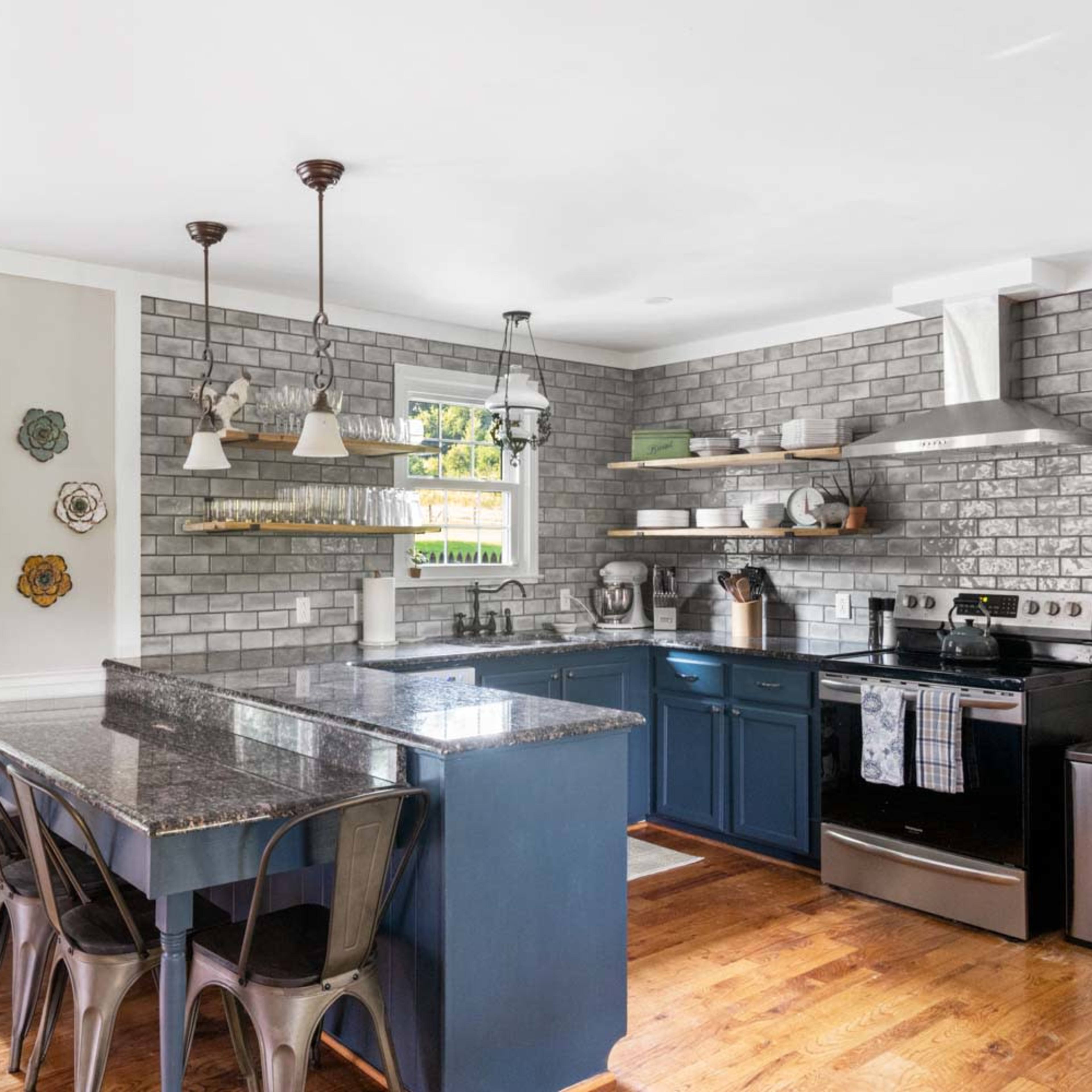 The kitchen features blue cabinets, gray tiled walls, and a central island with metal stools, illuminated by hanging light fixtures.