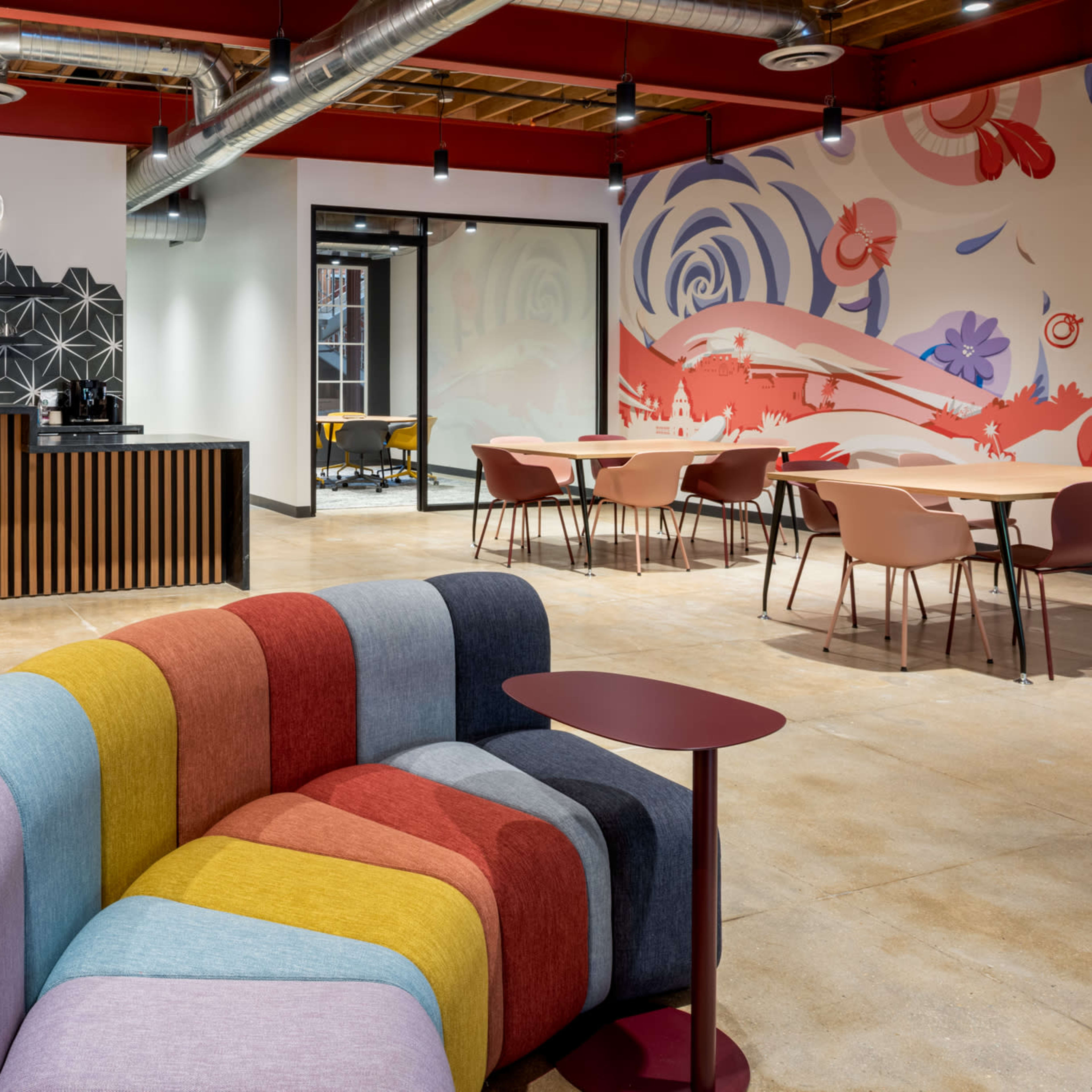 A modern office space featuring a colorful curved sofa, a bar-style counter with stools, and tables arranged for casual seating.