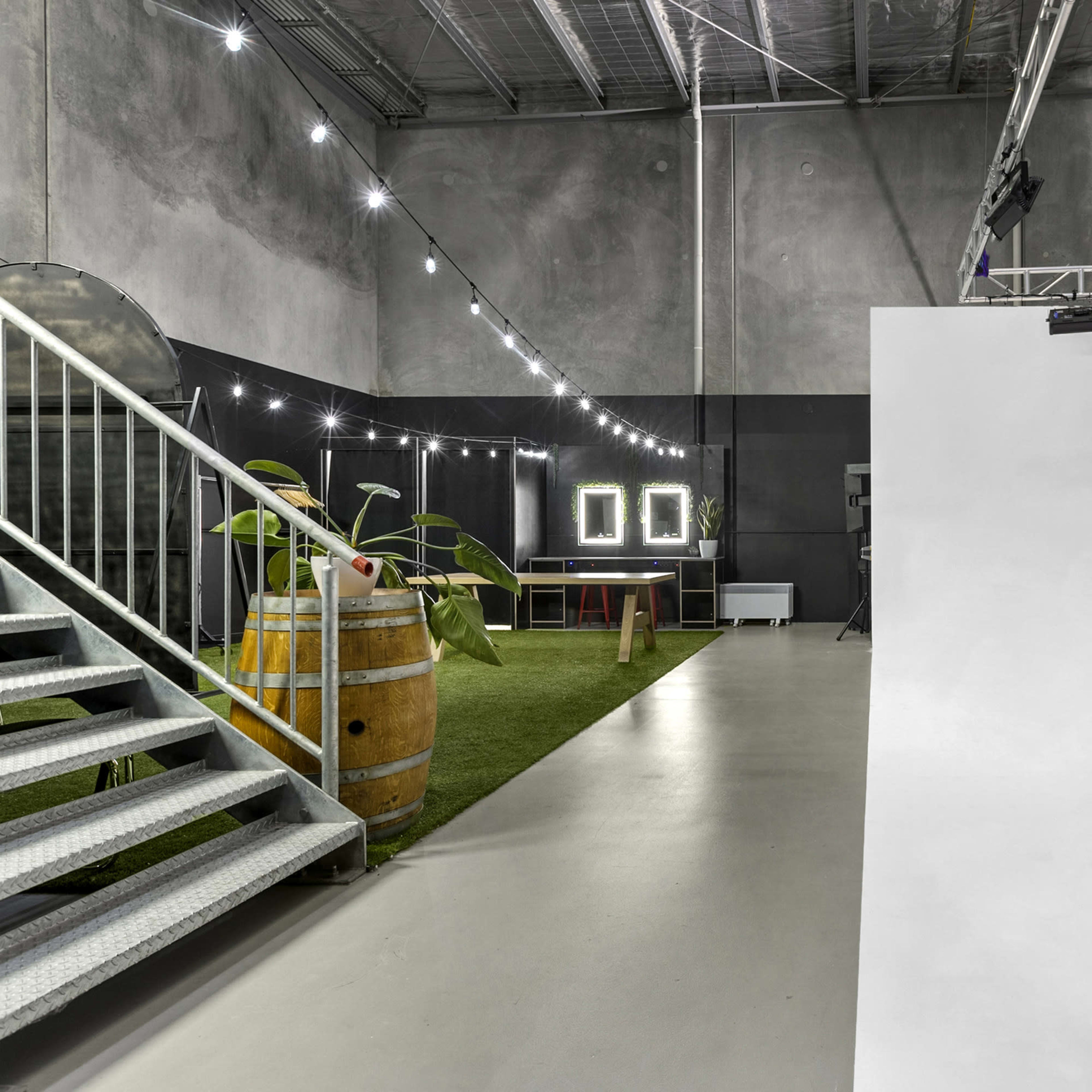 A spacious studio featuring a large empty white backdrop, artificial lighting, a wooden barrel, potted plants, and a set of framed mirrors on the wall.