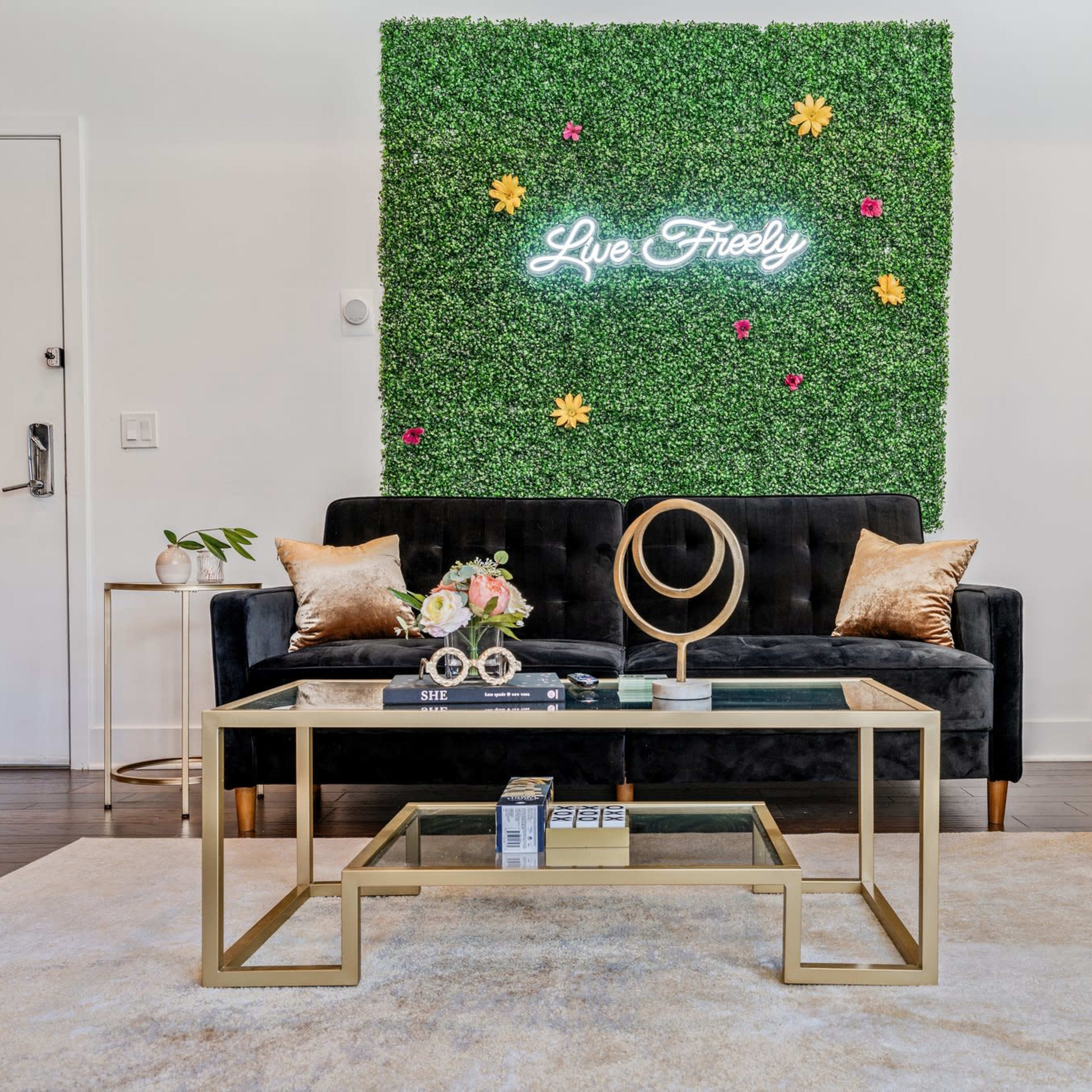 A cozy living room features a green wall with a neon sign reading "Live Freely," a black sofa, a glass coffee table, and a green accent chair.