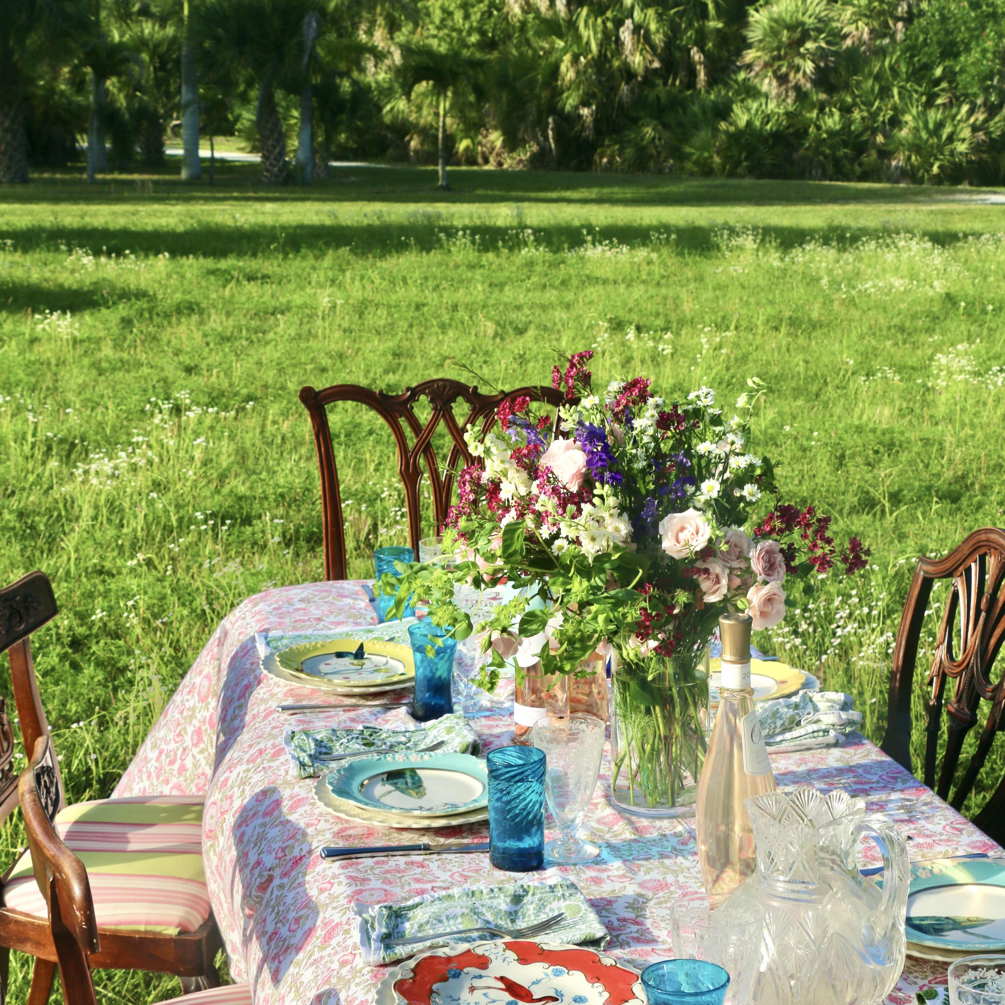 A table is set for a meal outdoors, adorned with colorful dishes, blue glasses, and a floral centerpiece, surrounded by a grassy area.