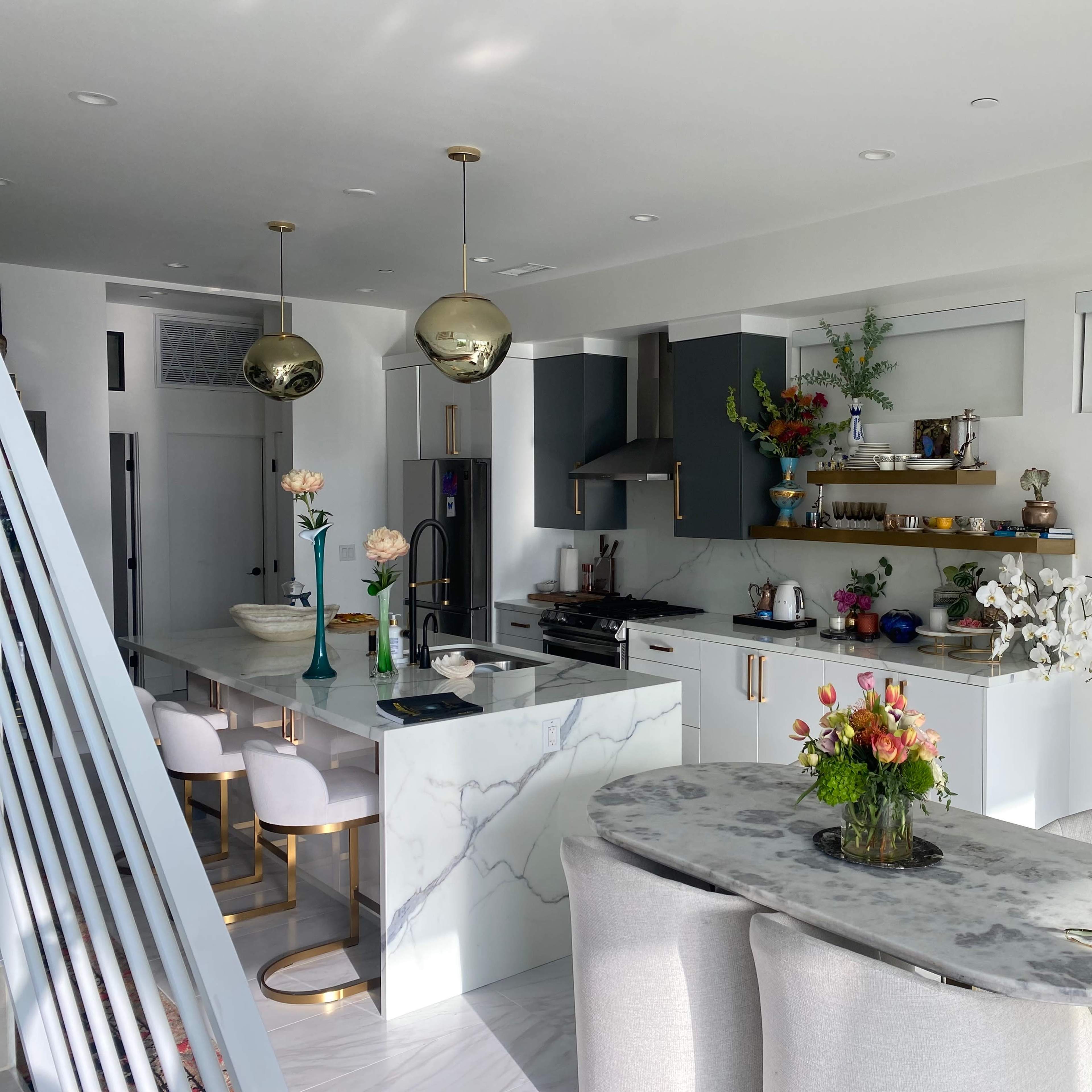A modern kitchen with marble countertops, gold light fixtures, and a dining area featuring a round table and upholstered chairs.