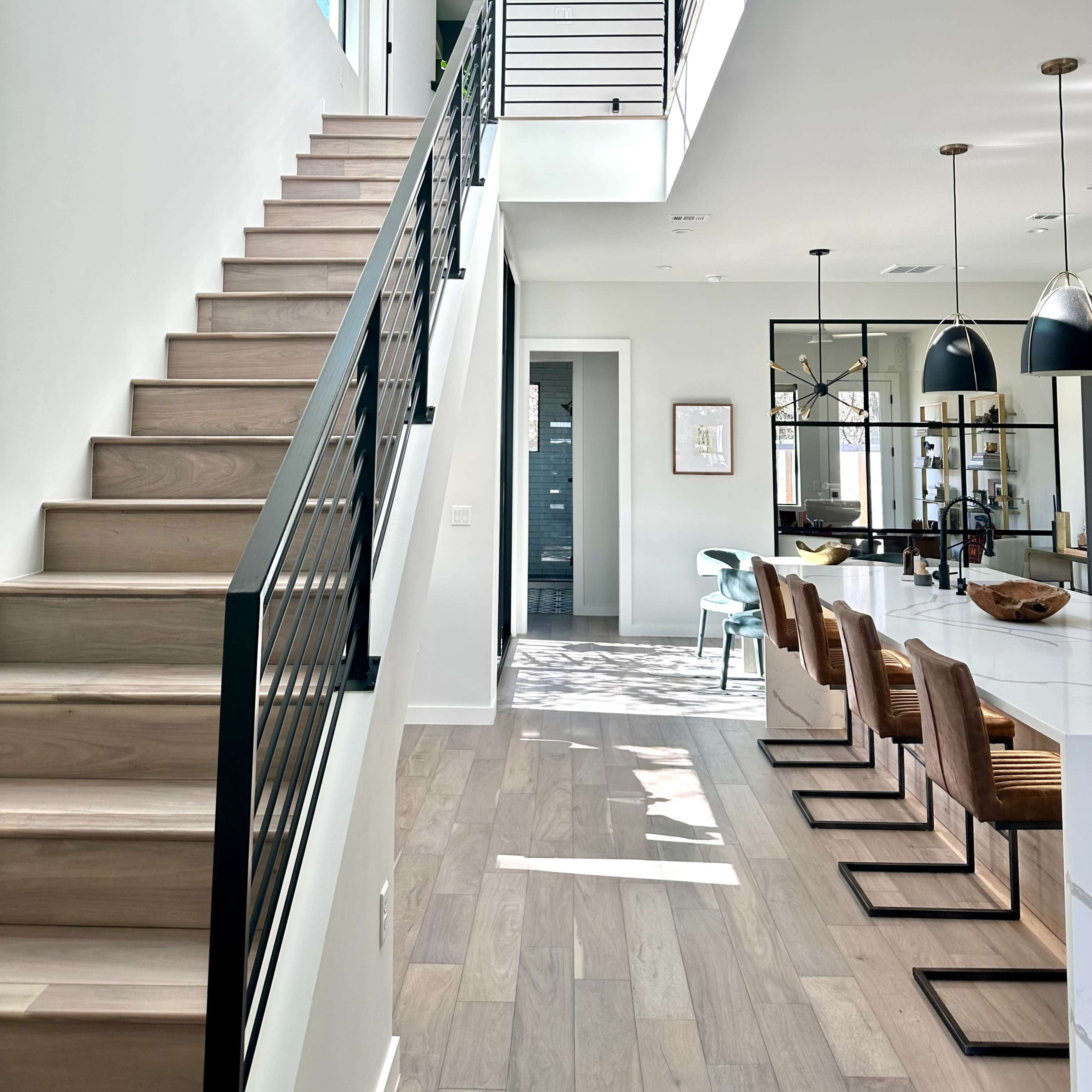 A staircase with sleek railings leads to an upper level in a modern home, featuring an open layout with a kitchen and dining area visible in the background.