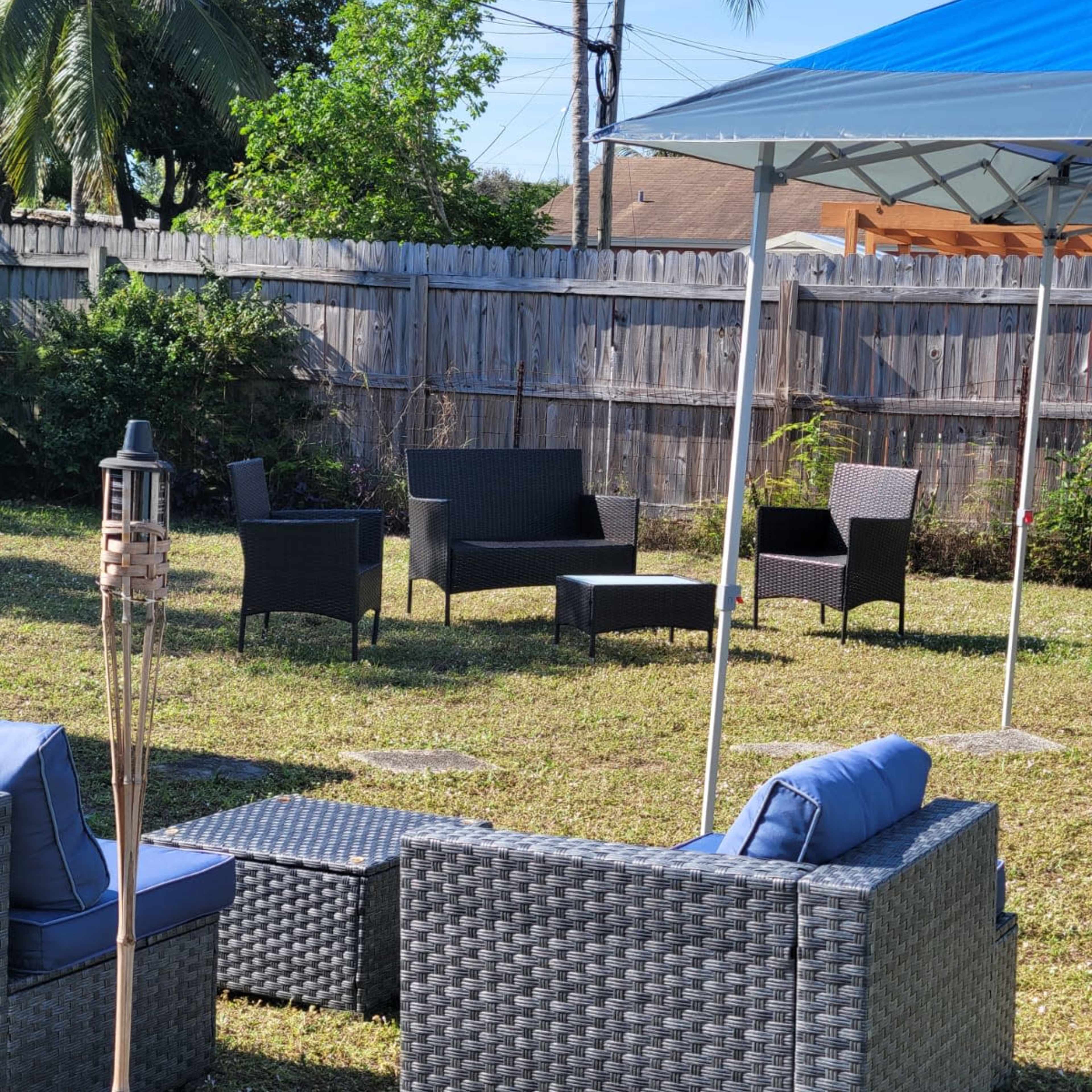 A backyard setup with several outdoor wicker chairs and tables arranged on green grass under a blue canopy.