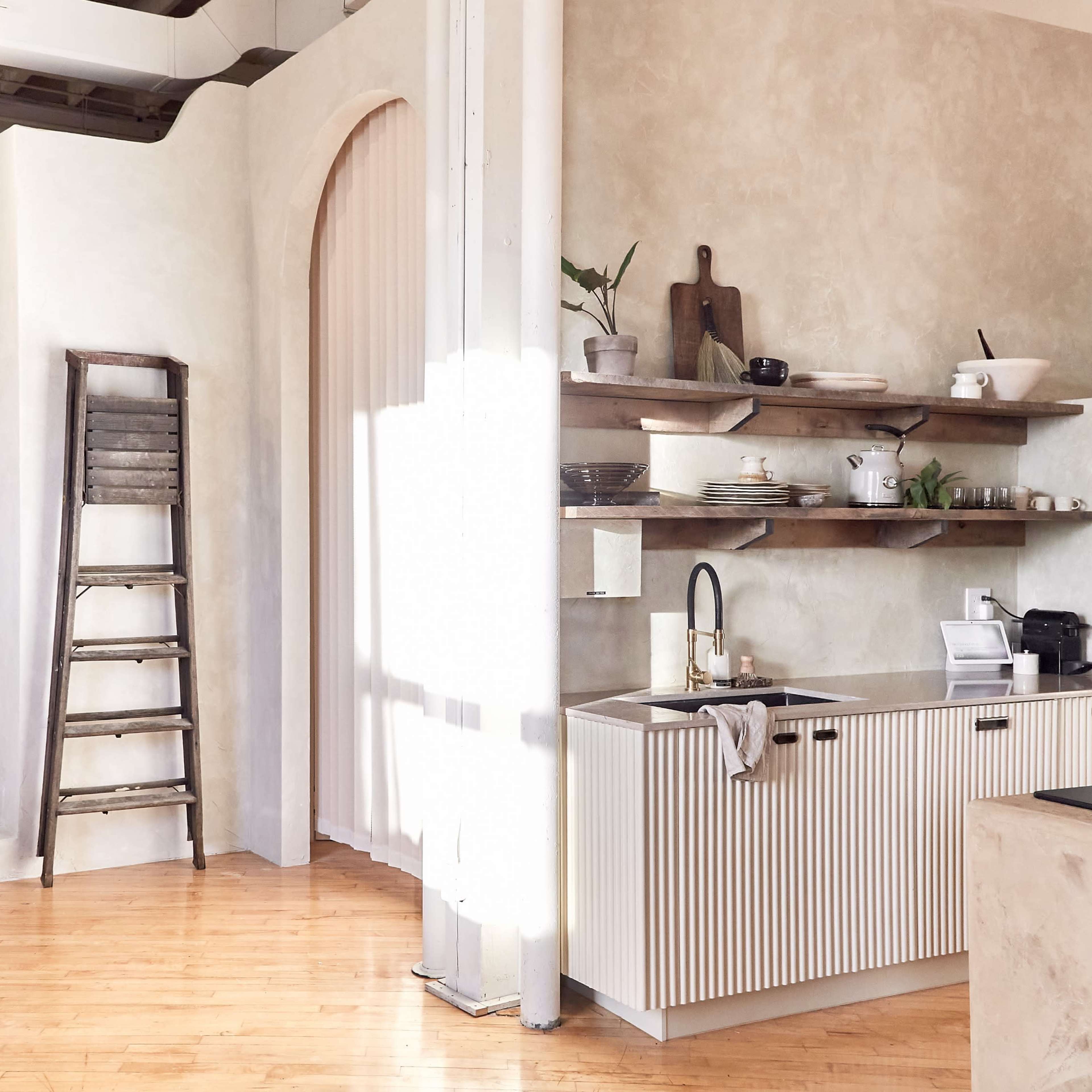 A minimalist interior space featuring a kitchen with a shelf and a ladder, alongside stairs leading to a different area with soft furniture.