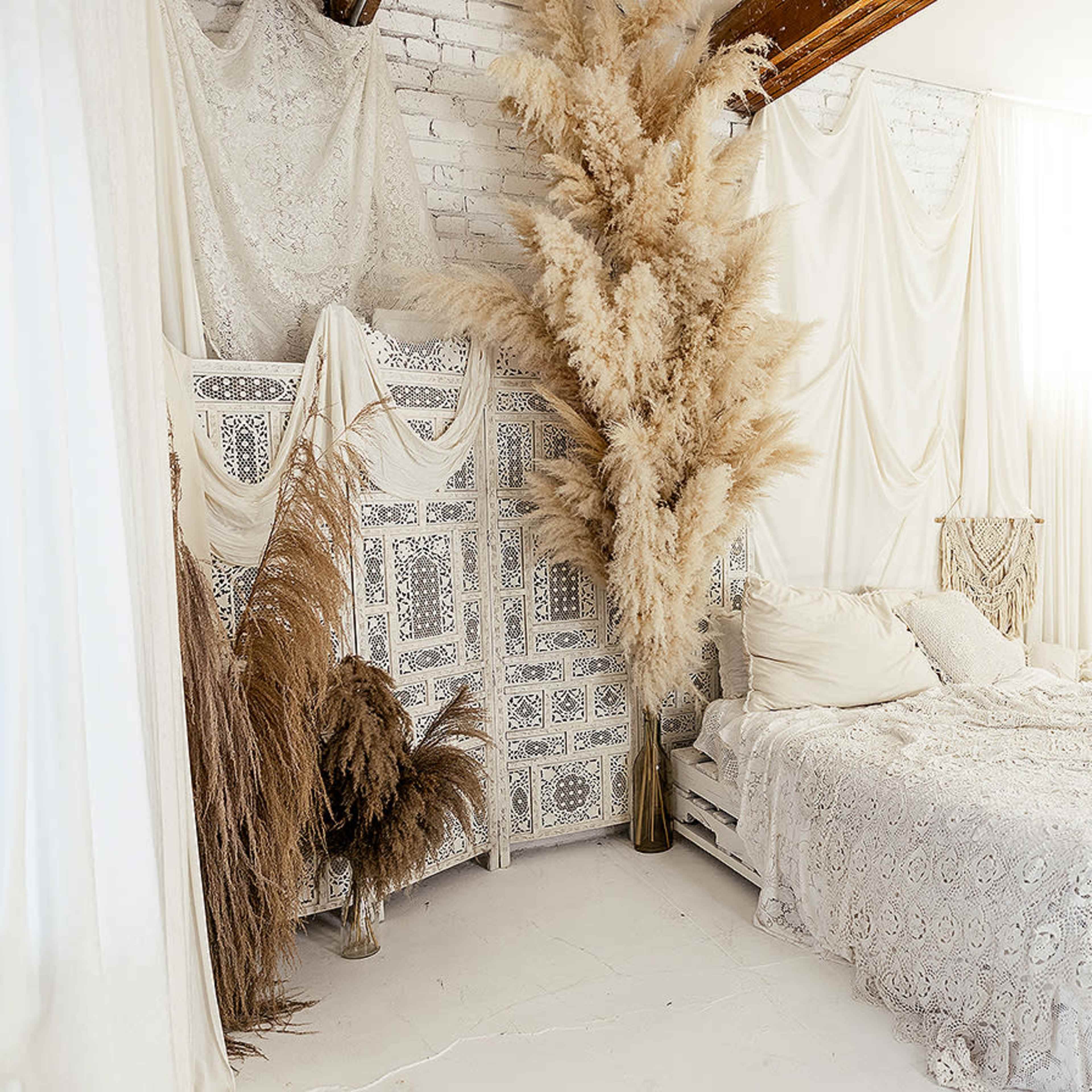 A cozy bedroom features a bed adorned with a lace coverlet, surrounded by beige plants and draped fabric against white brick walls.