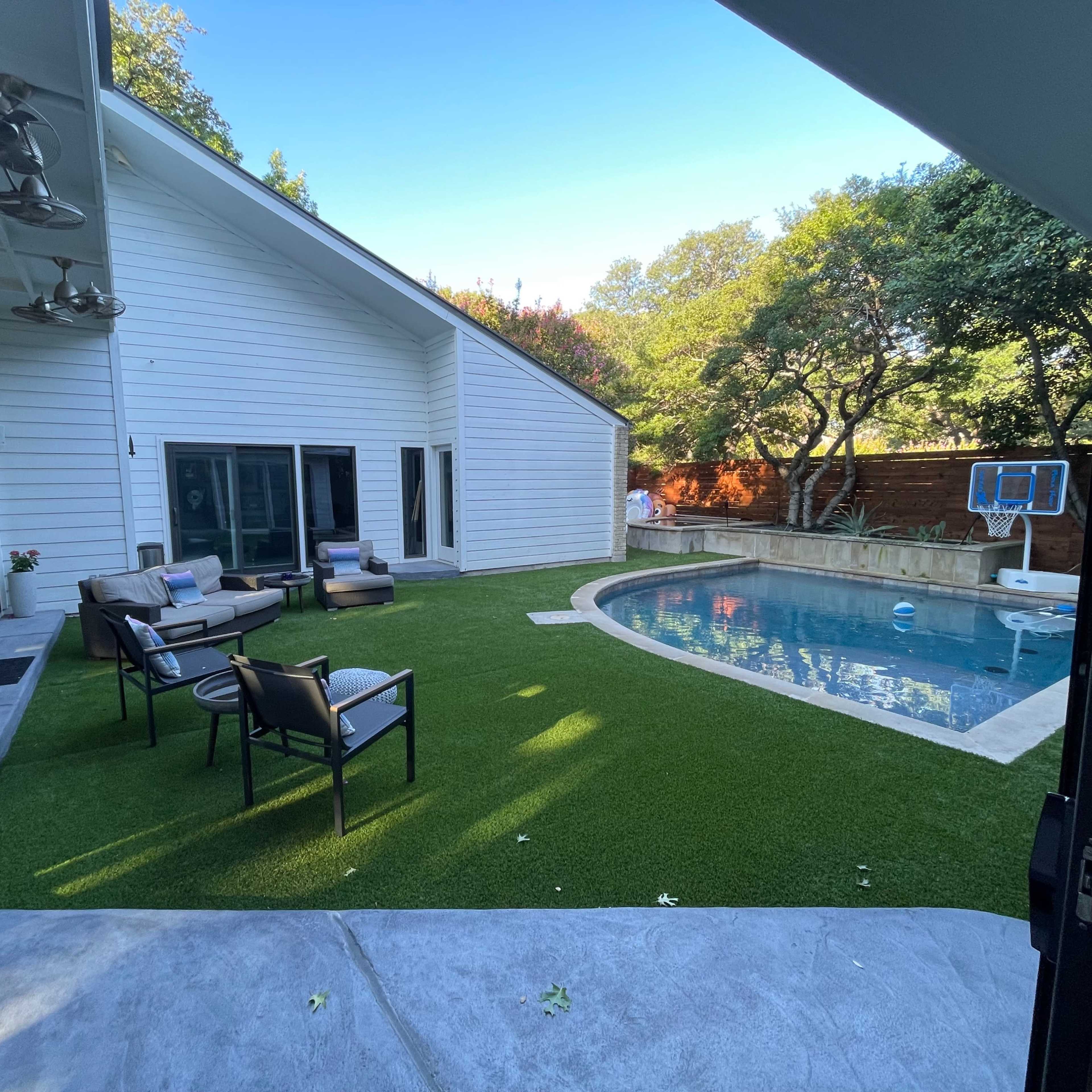 A modern backyard with a swimming pool, seating area, and artificial grass, surrounded by trees and a white house.
