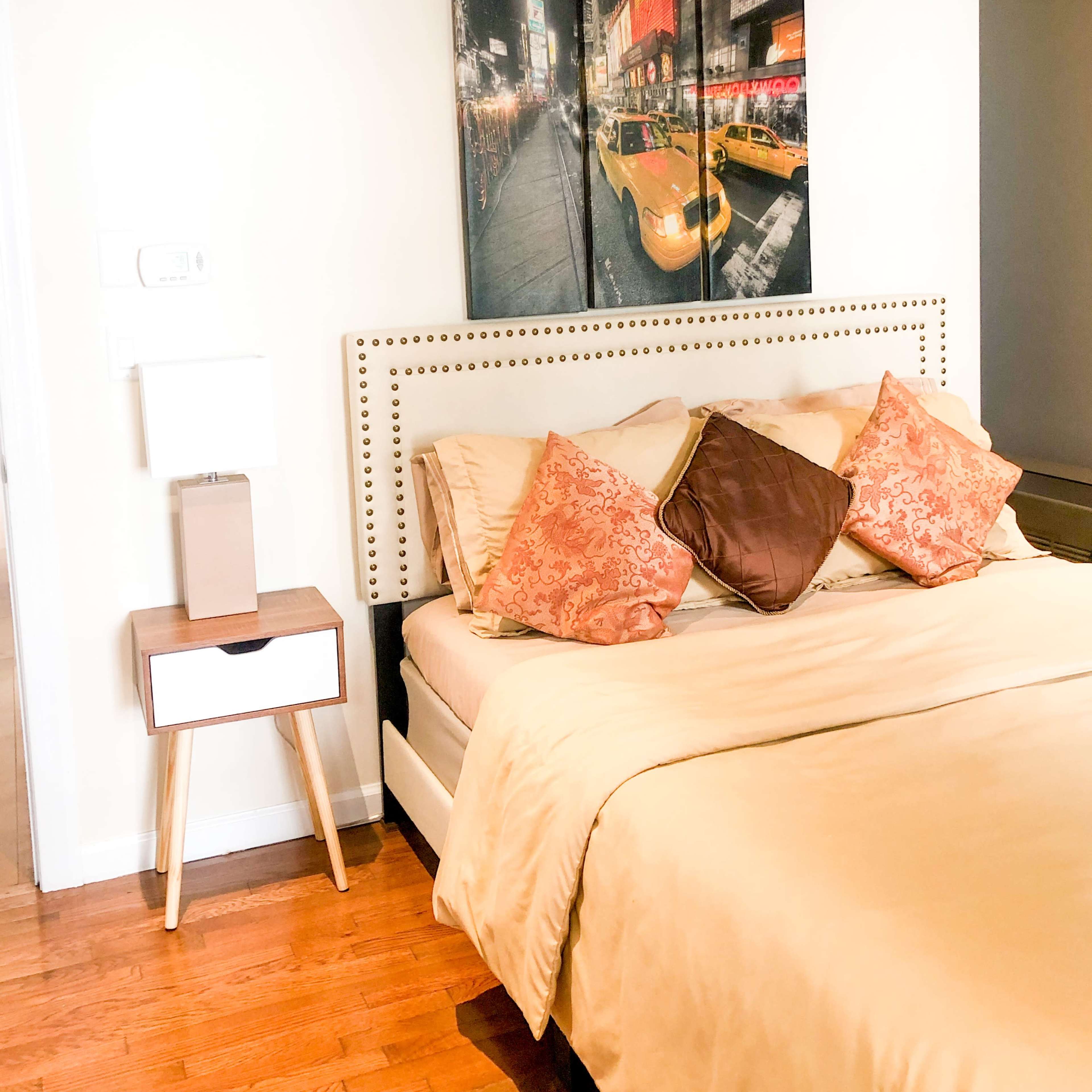 A bedroom with a bed featuring decorative pillows, a bedside table, and a large canvas print of a city scene with yellow taxis on the wall.