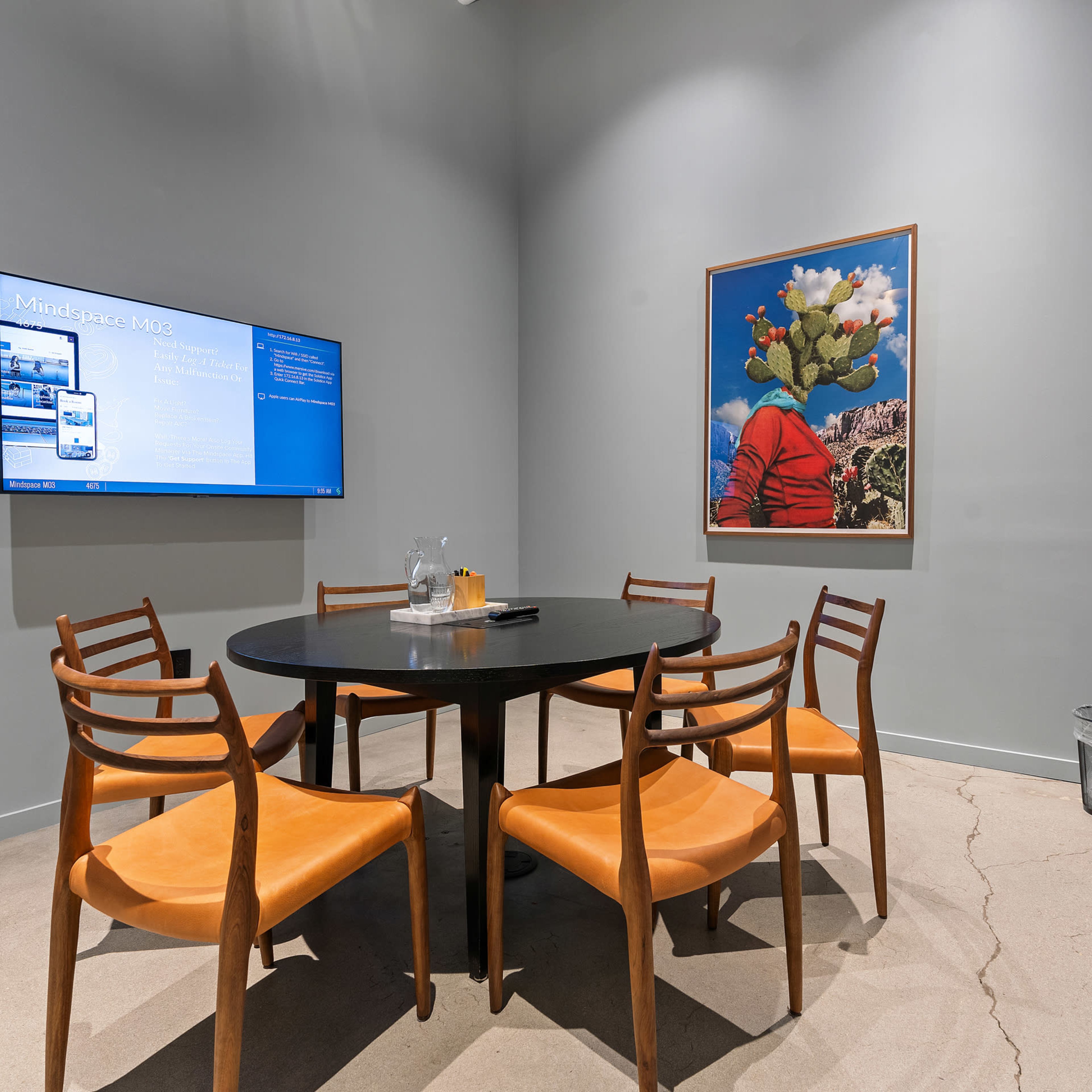 A modern meeting room features a round table surrounded by six wooden chairs, a wall-mounted screen displaying information, a framed artwork, and a coat rack in the corner.