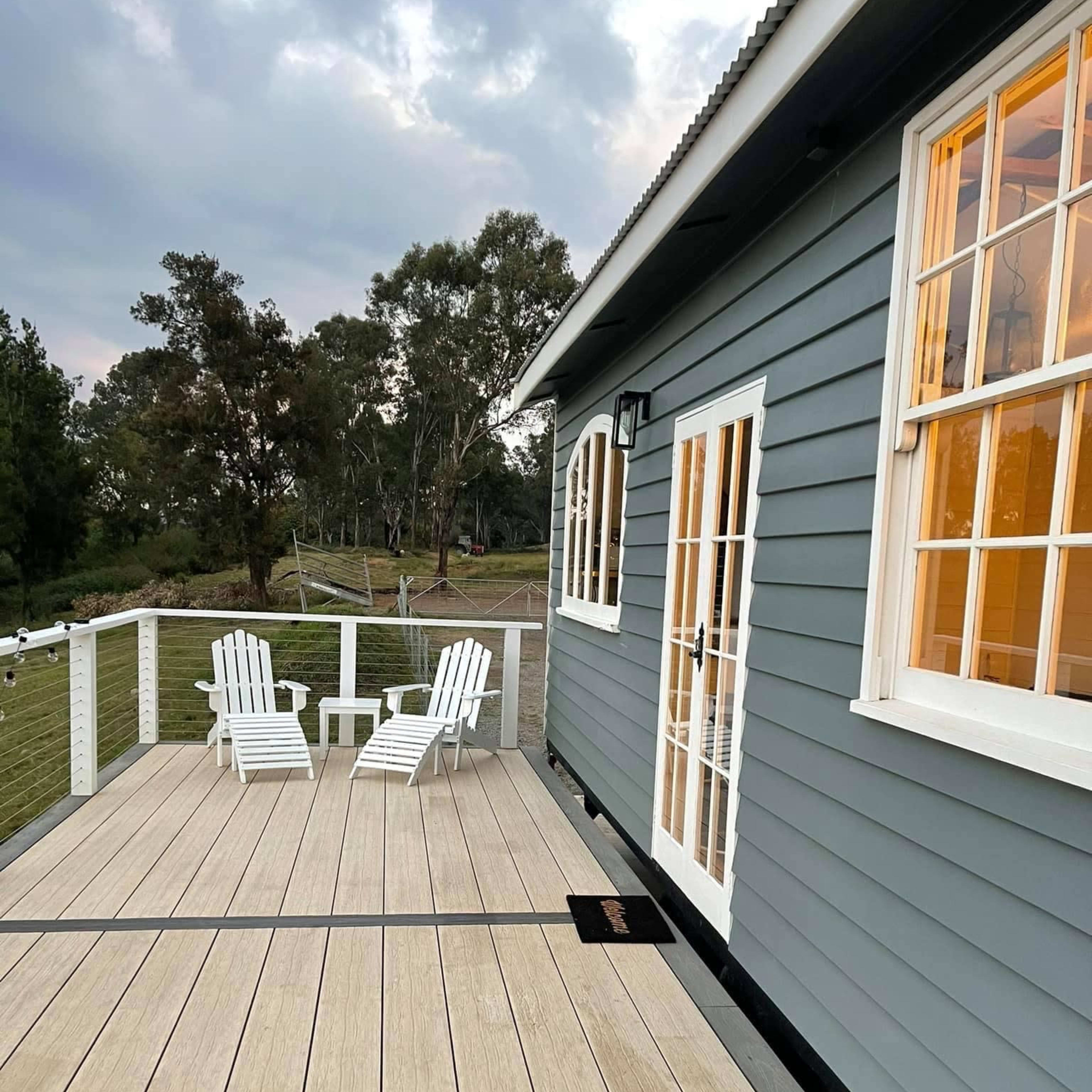 A wooden deck with two white chairs in front of a blue-gray house, and trees are visible in the background.